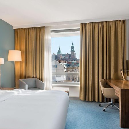 Radisson Blu Hotel Cracovia Esterno foto The photo depicts a hotel room featuring a large bed with white linens, a light-colored headboard, and a side lamp. There is a comfortable armchair in the corner, and a wooden desk with a chair positioned near the window. The window reveals a view of