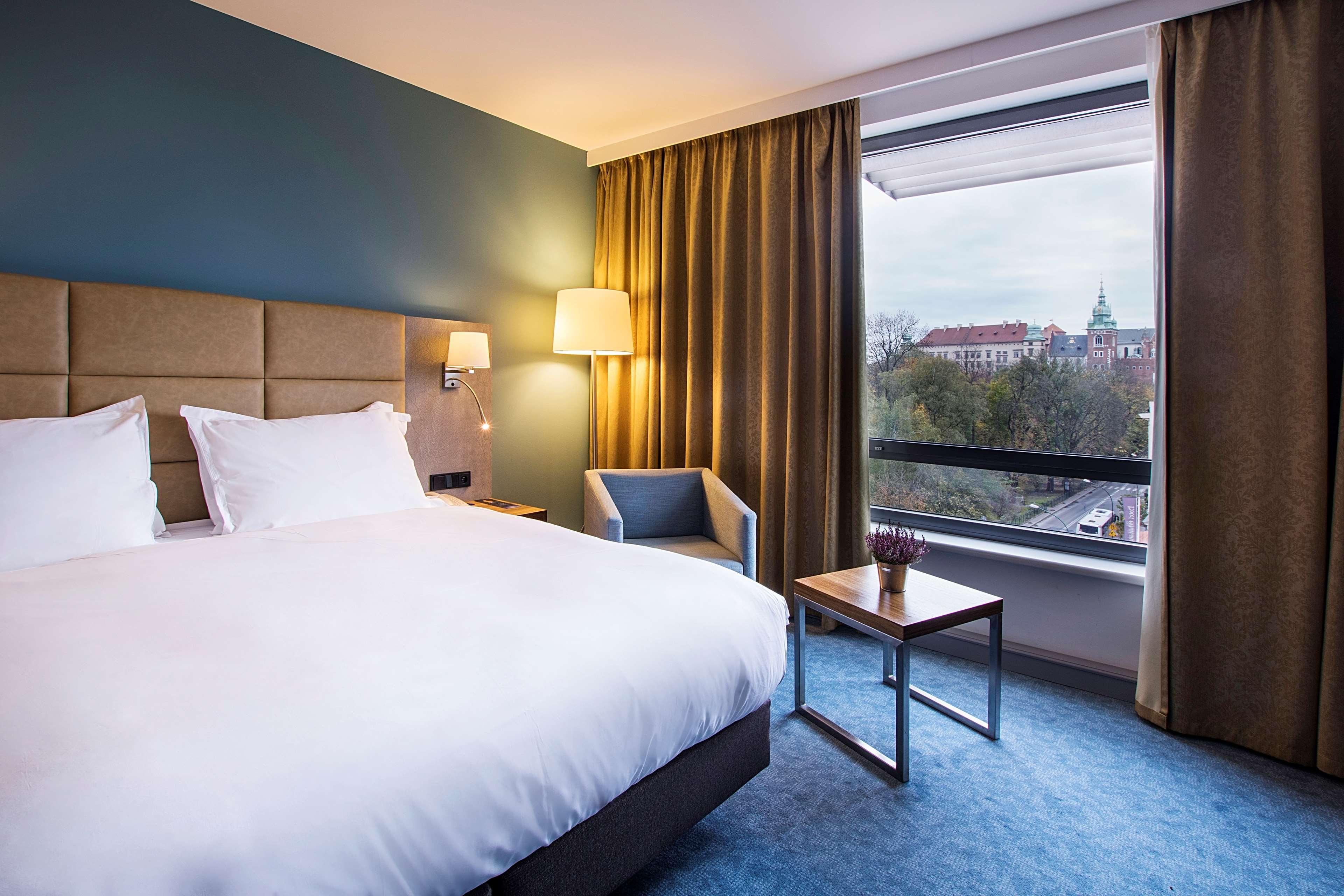 Radisson Blu Hotel Cracovia Esterno foto The photo shows a modern hotel room featuring a queen-size bed with white bedding and two pillows. There is a bedside lamp and a small armchair in the corner next to a window. The window has grey curtains, partially open, revealing a view of an exter