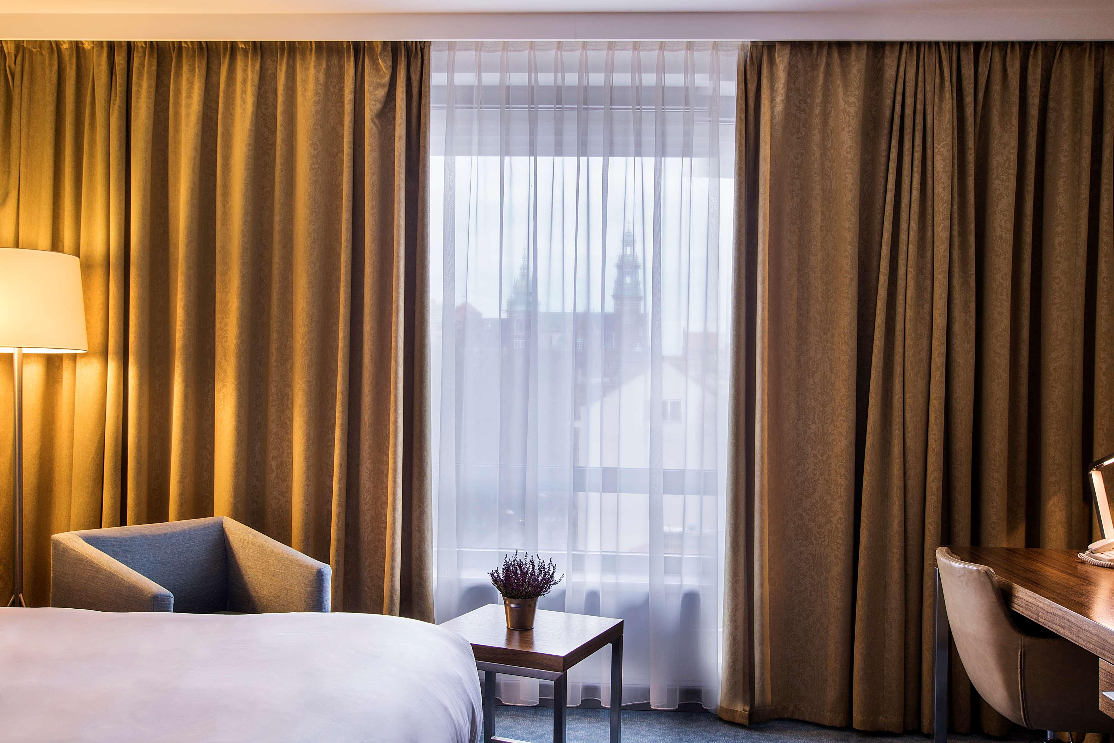 Radisson Blu Hotel Cracovia Esterno foto The photo shows a modern hotel room with a soft color palette. On the left, there is a comfortable armchair beside a small table with a decorative plant. The room has large windows covered with sheer curtains and gold-colored drapes, allowing soft li