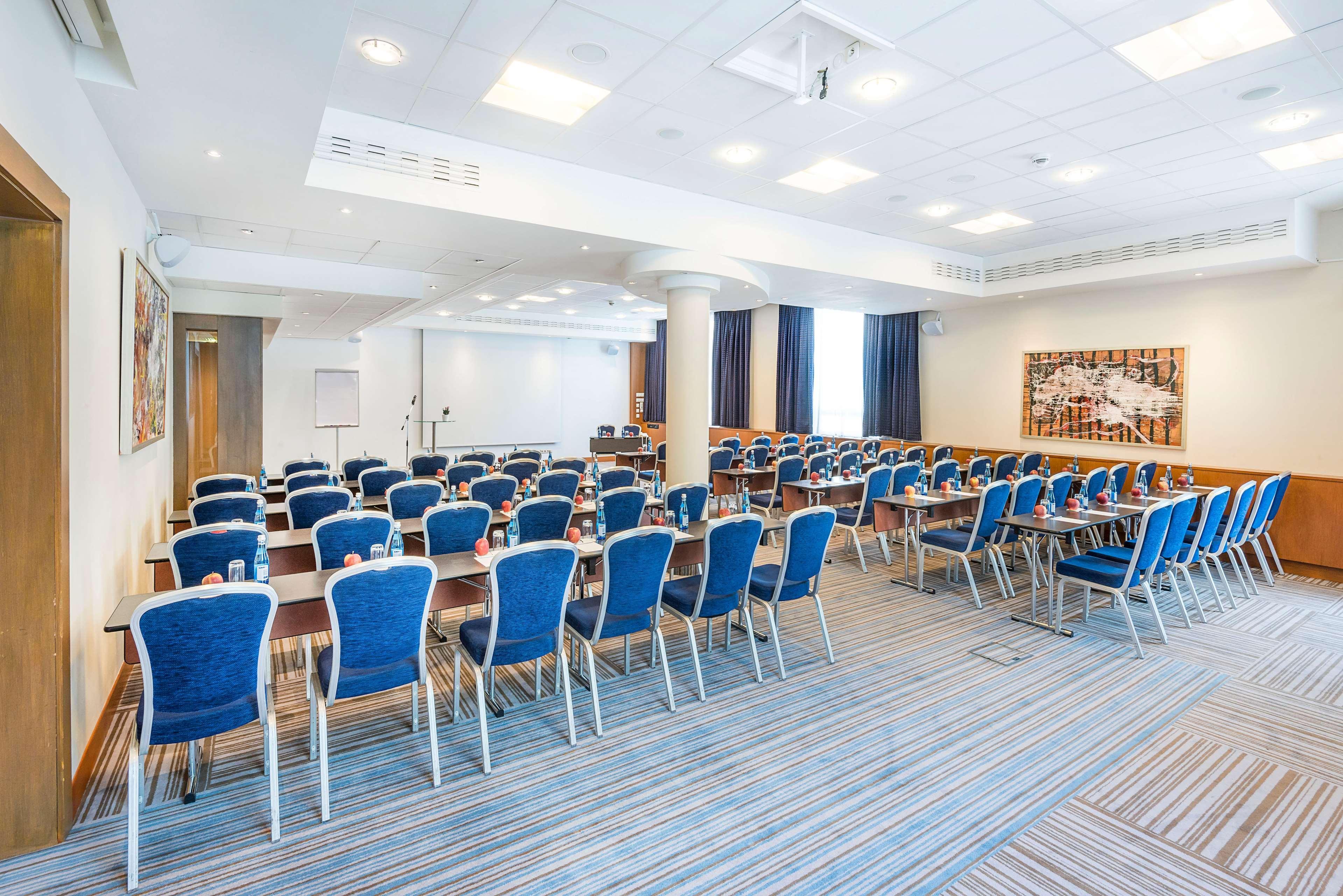 Radisson Blu Hotel Cracovia Esterno foto The photo shows a meeting or conference room set up for attendees. Rows of blue chairs are arranged around wooden tables, providing seating for participants. The room has a modern design with bright lighting, carpeting featuring a striped pattern, an