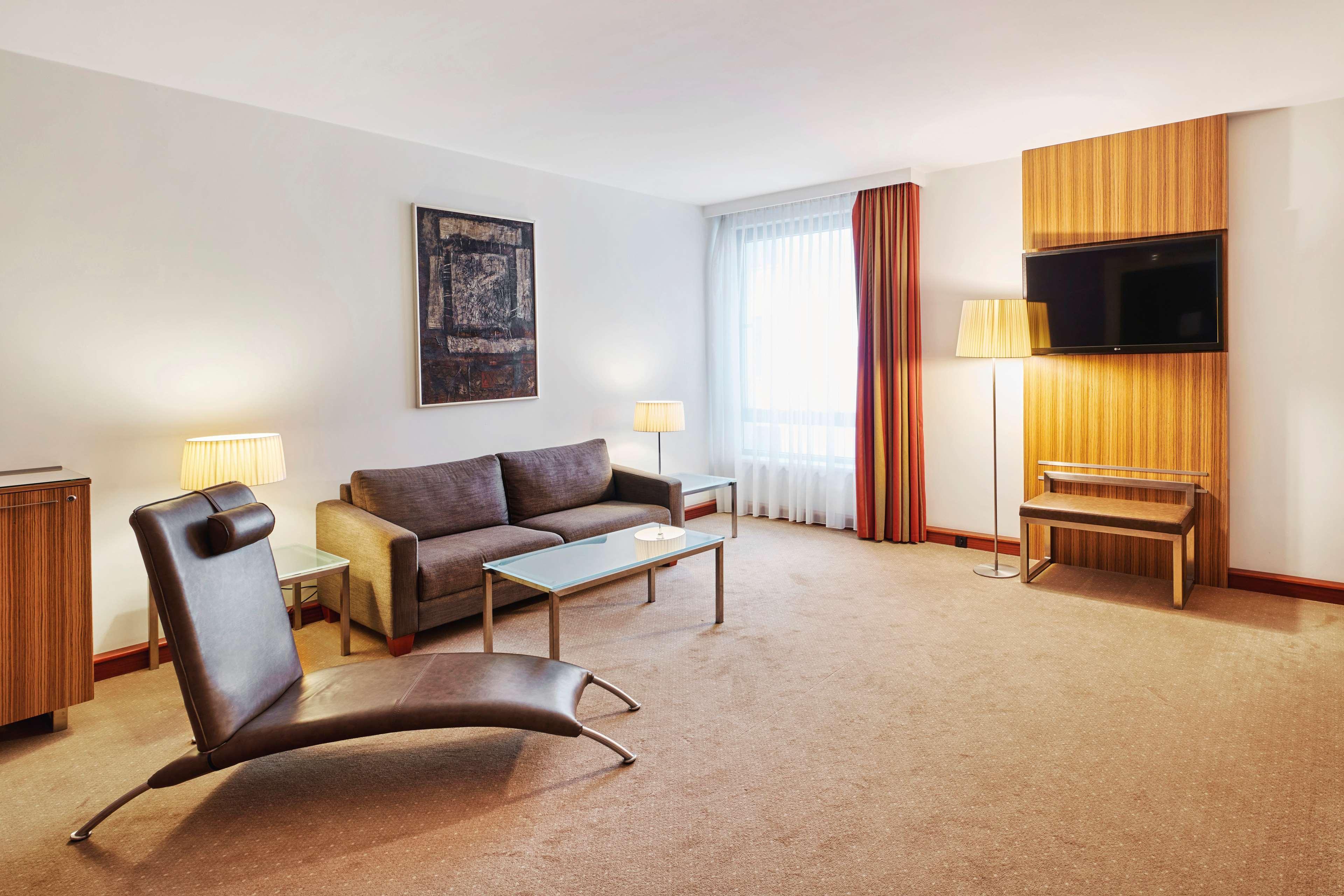 Radisson Blu Hotel Cracovia Esterno foto The photo shows a modern hotel room or lounge area. It features a comfortable gray sofa accompanied by a glass coffee table. There is a distinctive brown, stylish lounge chair, adding a unique design element to the space. The room is softly lit with 