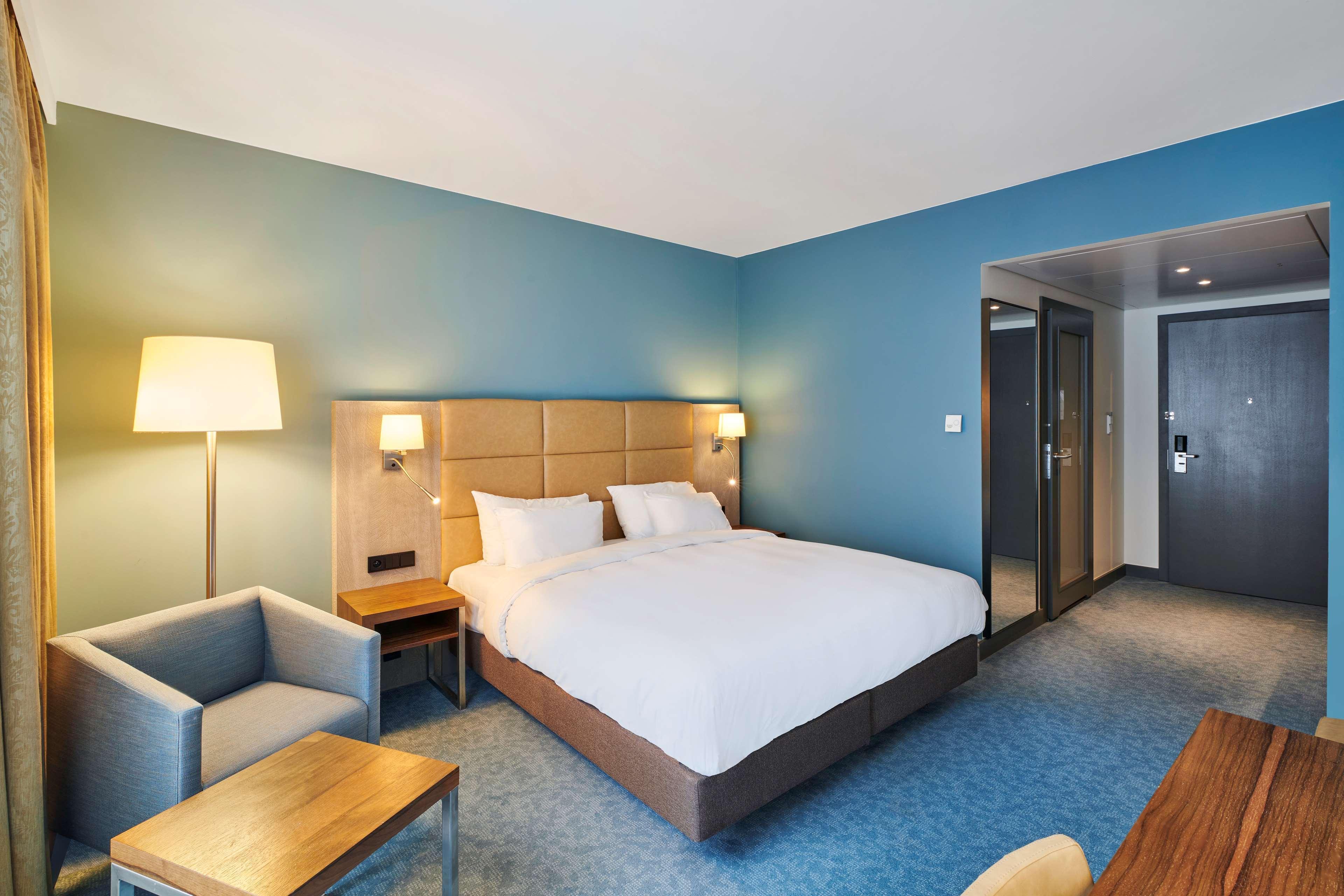 Radisson Blu Hotel Cracovia Esterno foto The photo shows a modern hotel room featuring a queen-sized bed with white linens and decorative pillows. There are bedside lamps on either side of the bed, and a light blue accent wall adds to the room's contemporary decor. To the side, there is a s