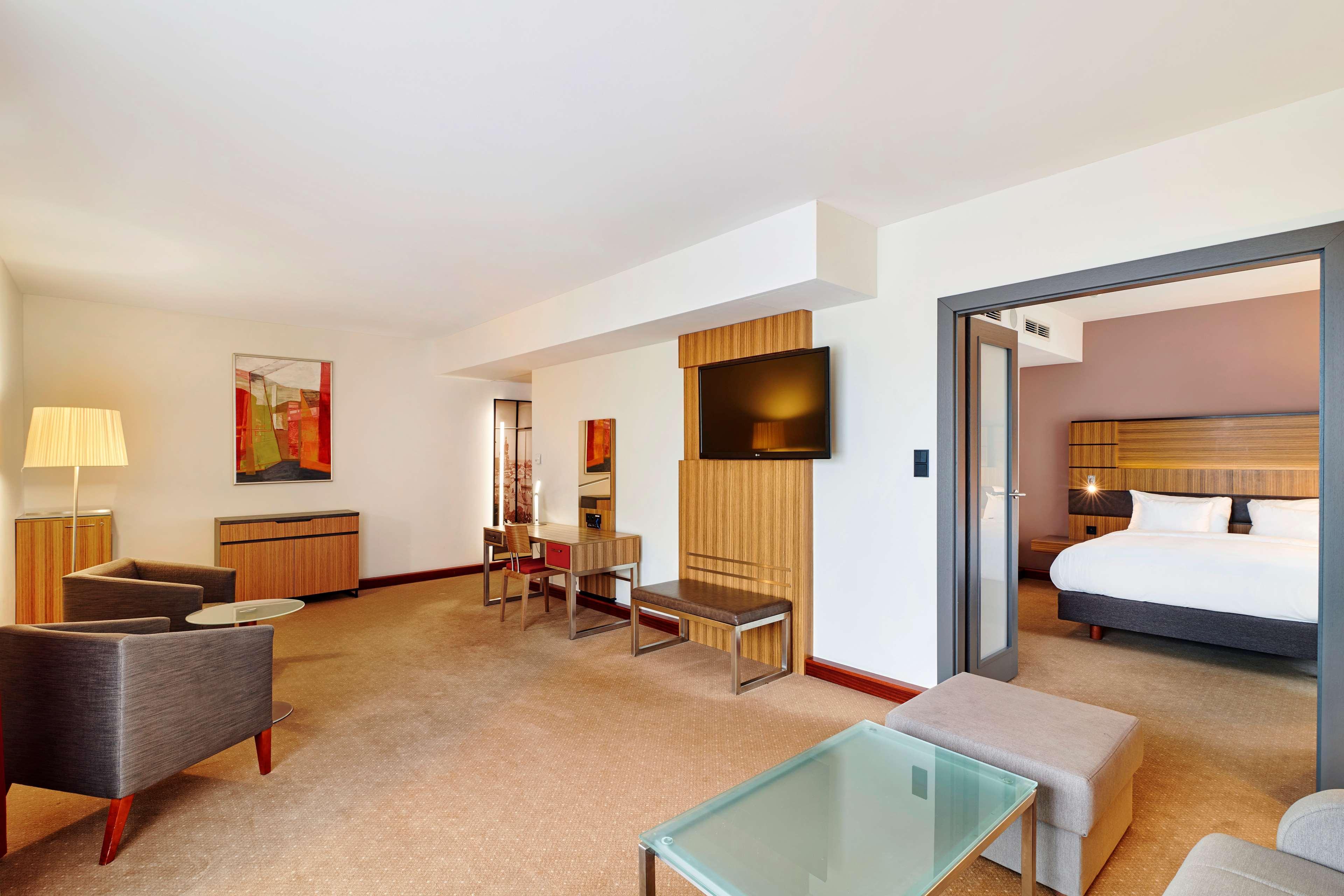 Radisson Blu Hotel Cracovia Esterno foto The photo shows a hotel room or suite that features a spacious and modern design. On one side, there are two gray armchairs accompanied by a small table, creating a cozy seating area. A glass-top table and some additional seating can also be seen.

I