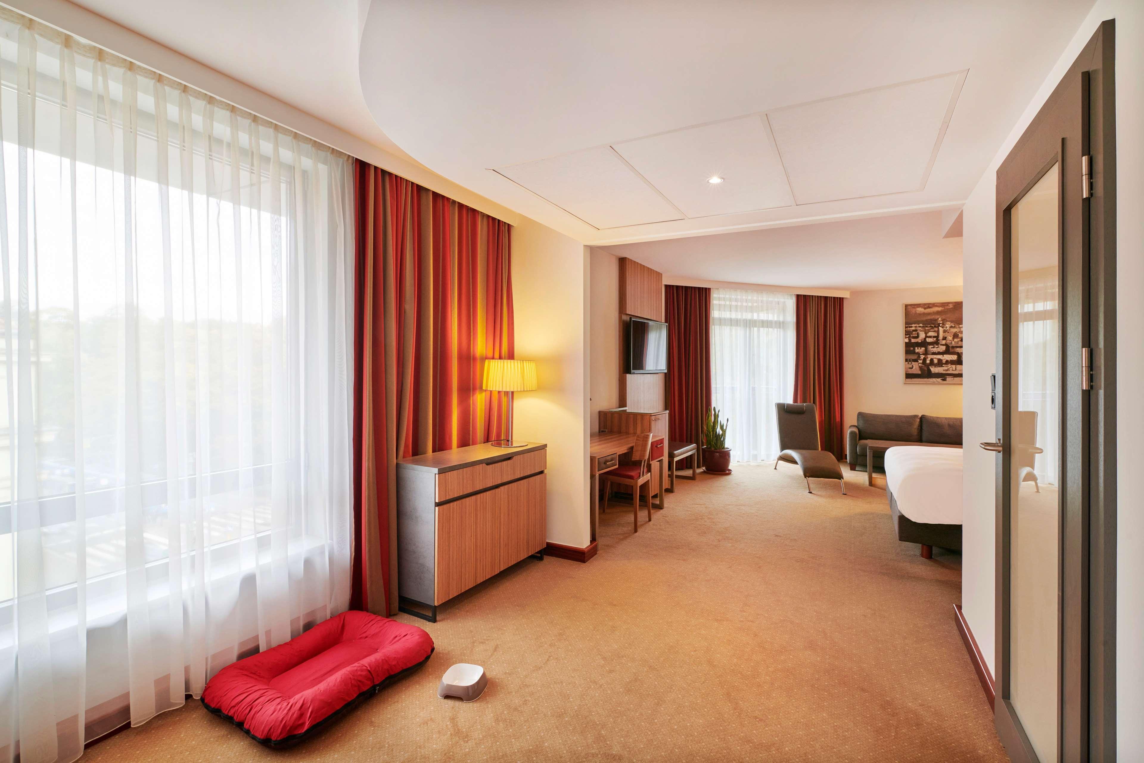 Radisson Blu Hotel Cracovia Esterno foto The photo shows a hotel room interior that features an inviting and modern design. On one side, there is a large window with sheer curtains allowing natural light to fill the space. The walls are adorned with warm tones, complemented by rich red curt