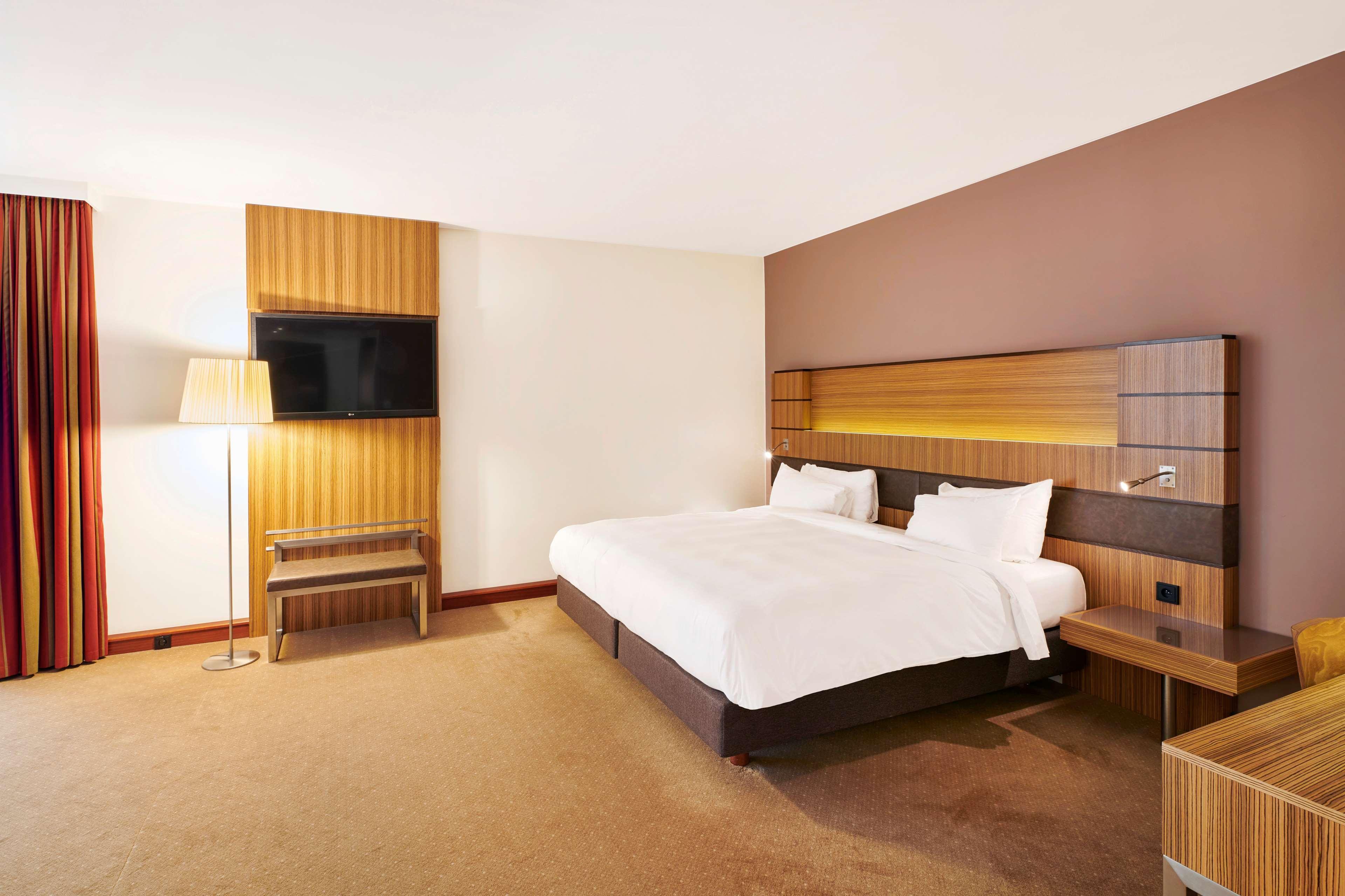 Radisson Blu Hotel Cracovia Esterno foto The photo shows a modern hotel room featuring a large double bed with white bedding and pillows. There is a wooden headboard with integrated lighting above the bed. On the wall opposite the bed, a flat-screen television is mounted. To the side, there