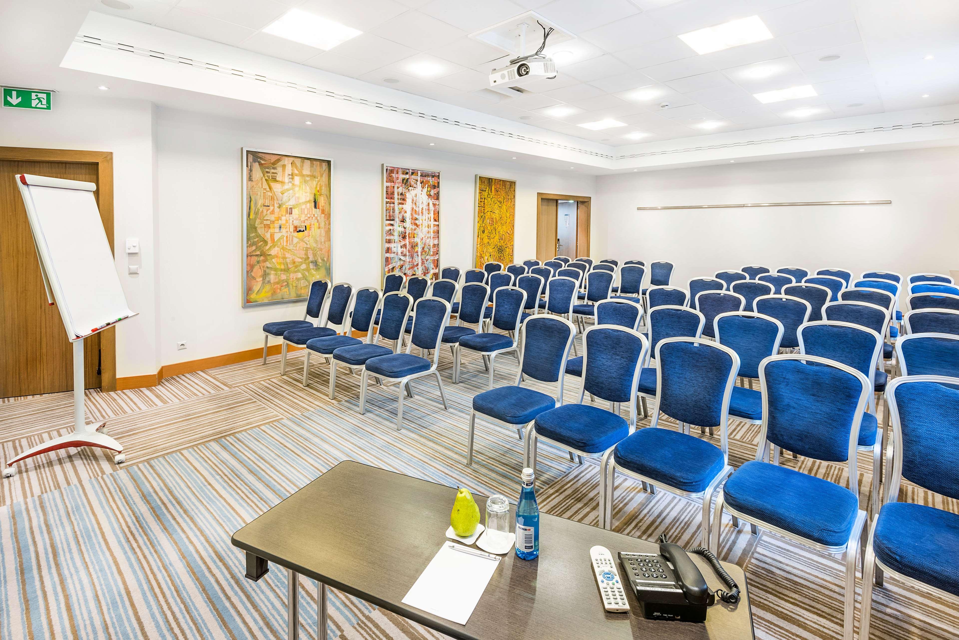Radisson Blu Hotel Cracovia Esterno foto The photo shows a conference room set up for an event or meeting. There are multiple rows of blue chairs arranged in a semi-theater style, facing a presentation area. In the foreground, there is a table with a few items, including a bottle of water a