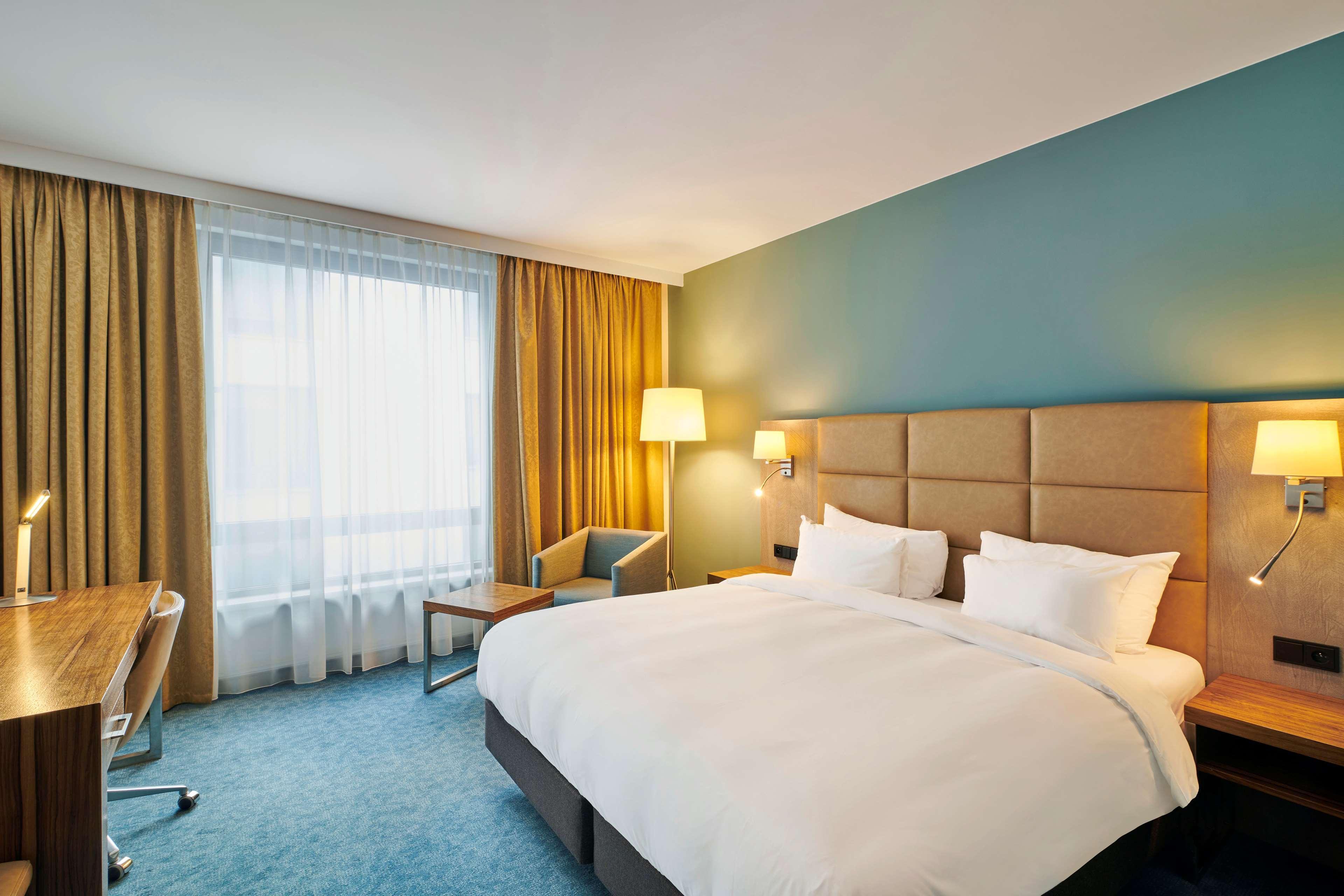Radisson Blu Hotel Cracovia Esterno foto The photo shows a modern hotel room. It features a large bed with white bedding and pillows. The walls are painted a calming blue, and there are large windows covered with sheer curtains, allowing natural light to enter. Next to the bed, there are be