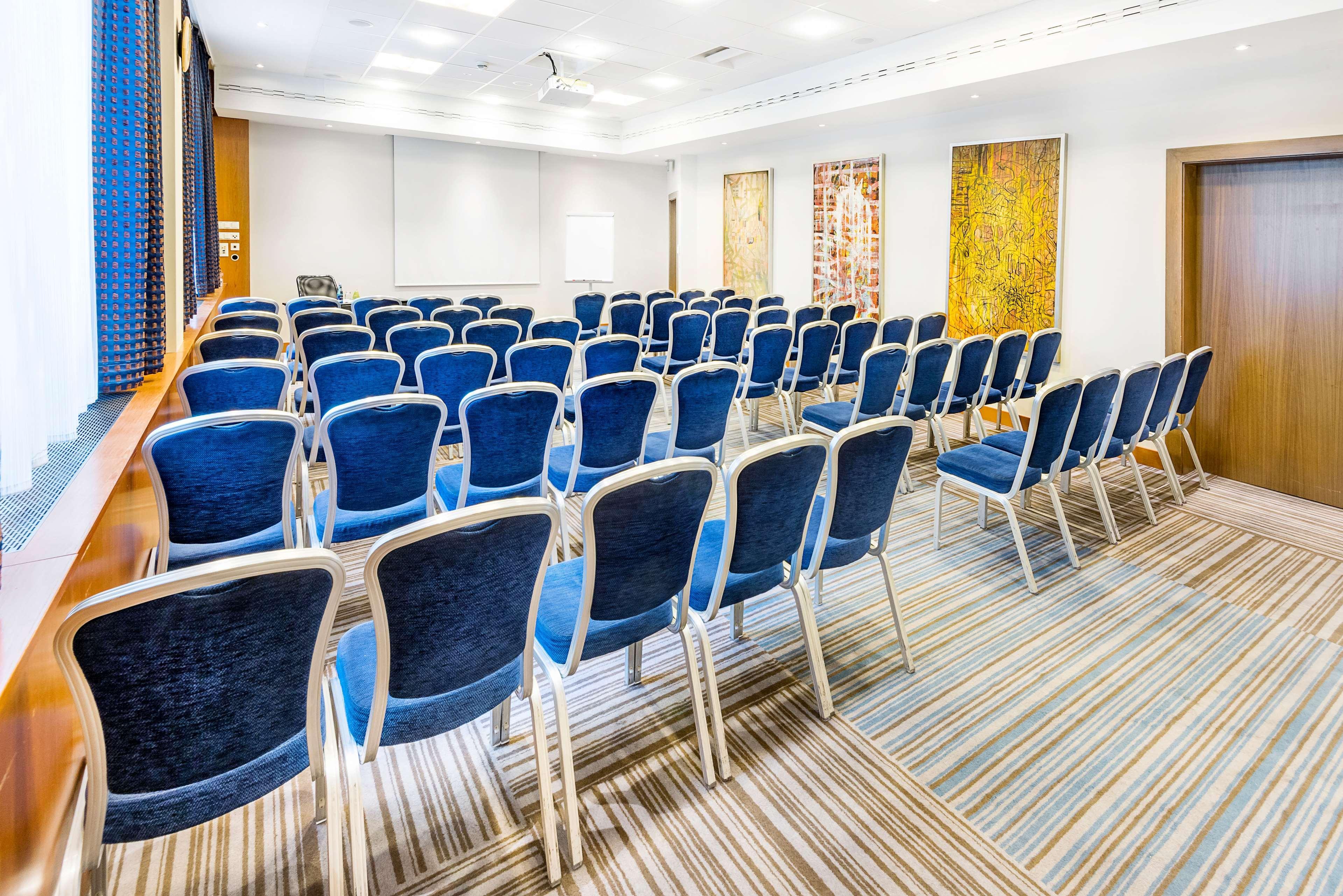 Radisson Blu Hotel Cracovia Esterno foto The photo shows an indoor conference room set up for a presentation or meeting. There are several rows of blue chairs arranged facing the front of the room. The chairs have silver metal frames and plush cushions. On the walls, there are decorative pa