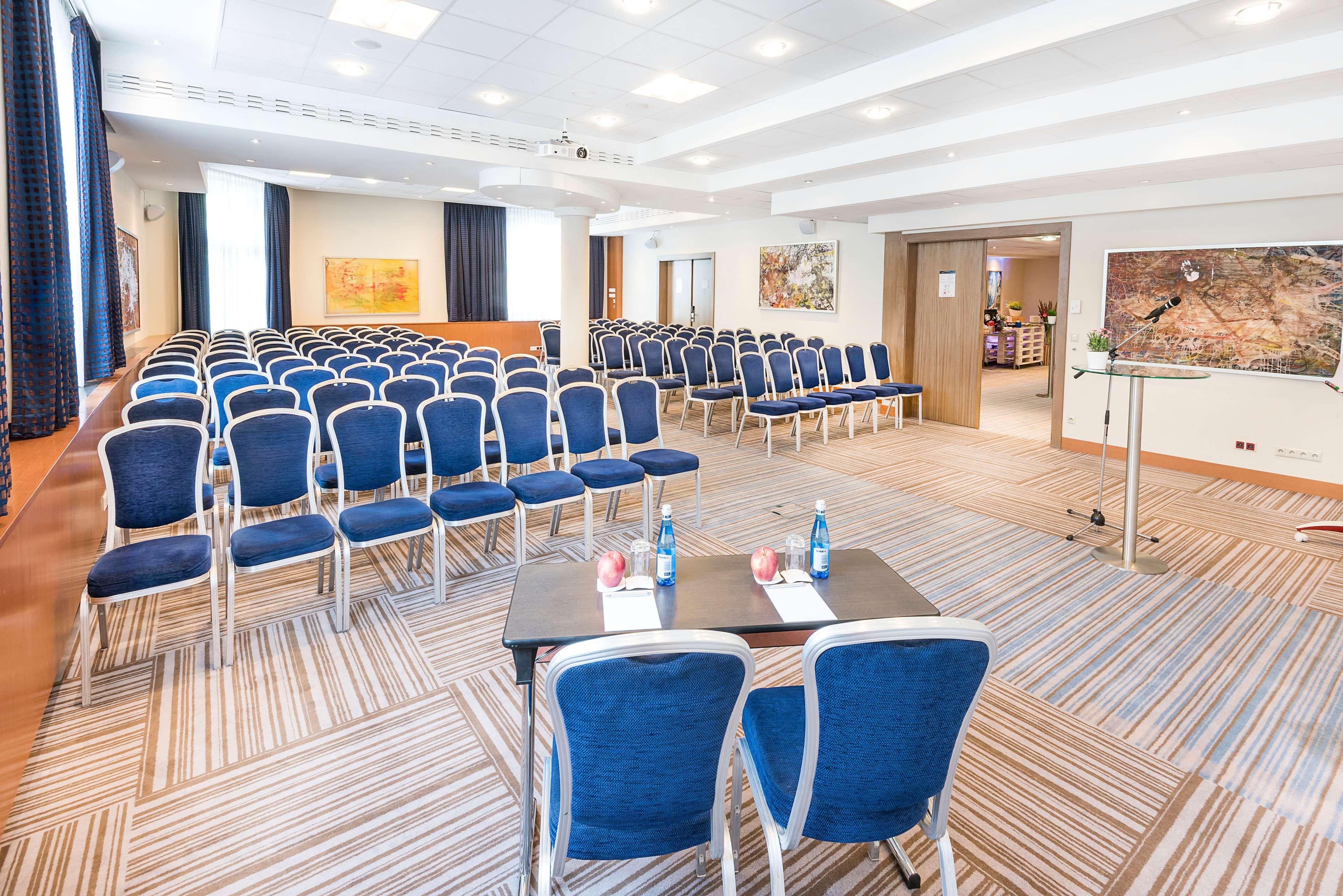 Radisson Blu Hotel Cracovia Esterno foto The photo shows a conference room set up for an event. There are several rows of blue upholstered chairs arranged facing a small stage or presentation area. In the foreground, there are two tables with water bottles and some red decorations on them. 