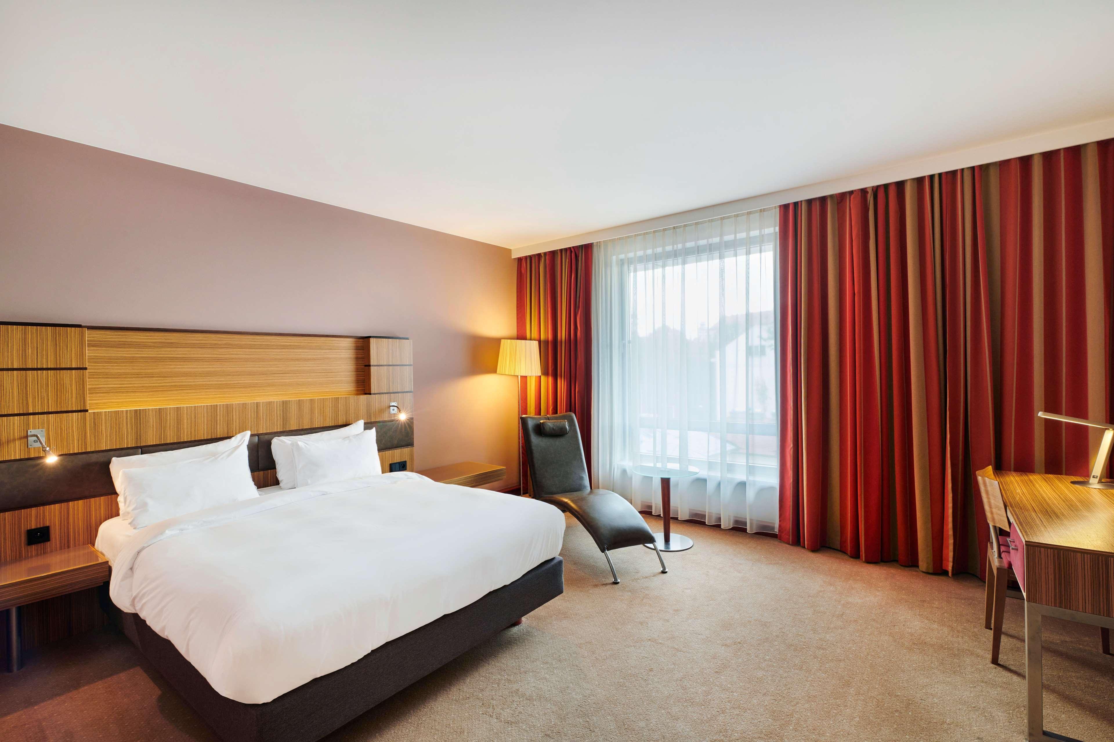 Radisson Blu Hotel Cracovia Esterno foto The photo shows a modern hotel room featuring a large bed with white bedding and two pillows. The walls are painted in a warm tone, and there's a stylish wooden headboard. Next to the bed, there's an elegant lamp providing soft lighting. A large wind