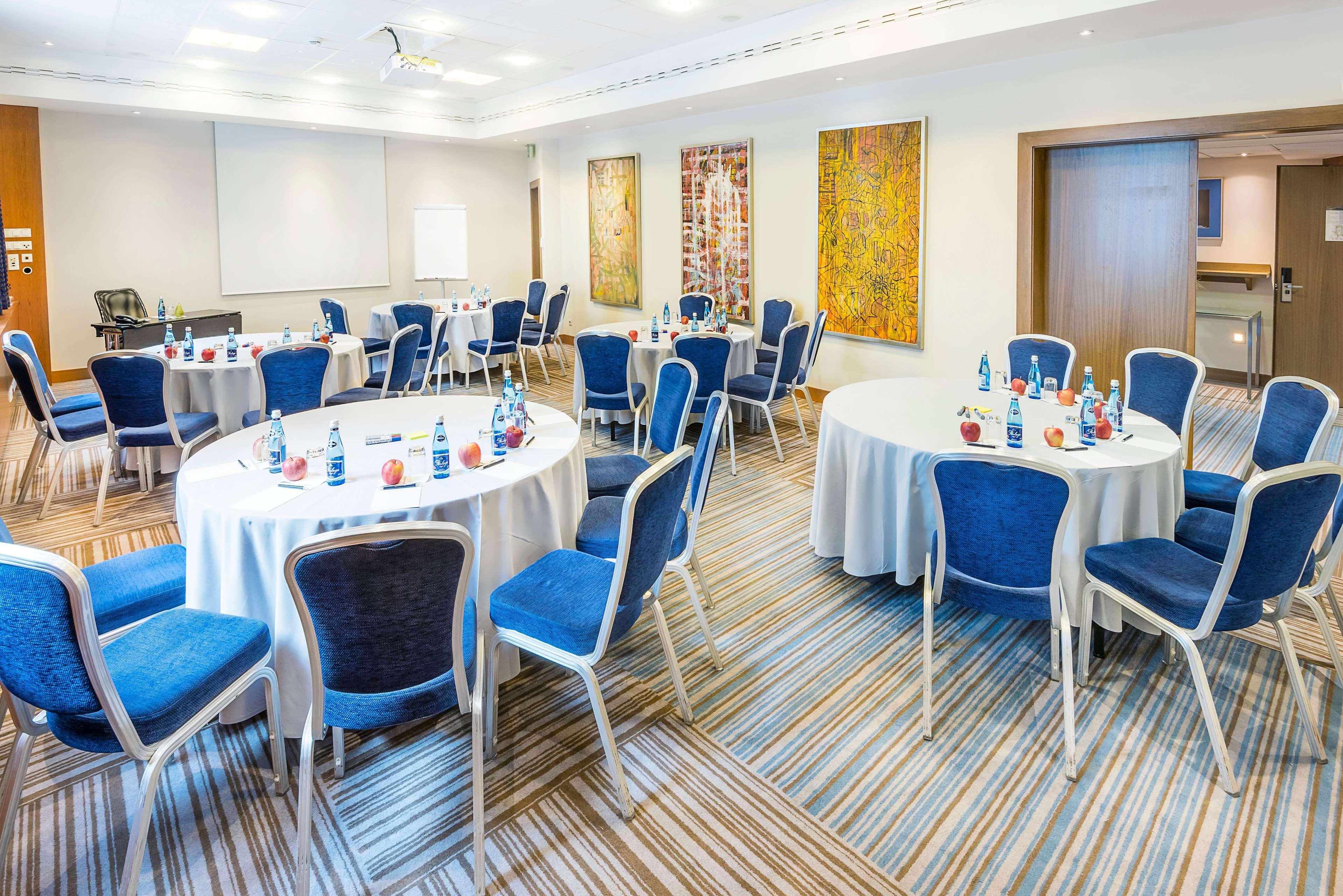 Radisson Blu Hotel Cracovia Esterno foto The photo shows a conference or meeting room set up for an event. There are several round tables covered with white tablecloths, each accompanied by blue chairs. Bottled water and small notepads appear to be placed on the tables. The room features mo