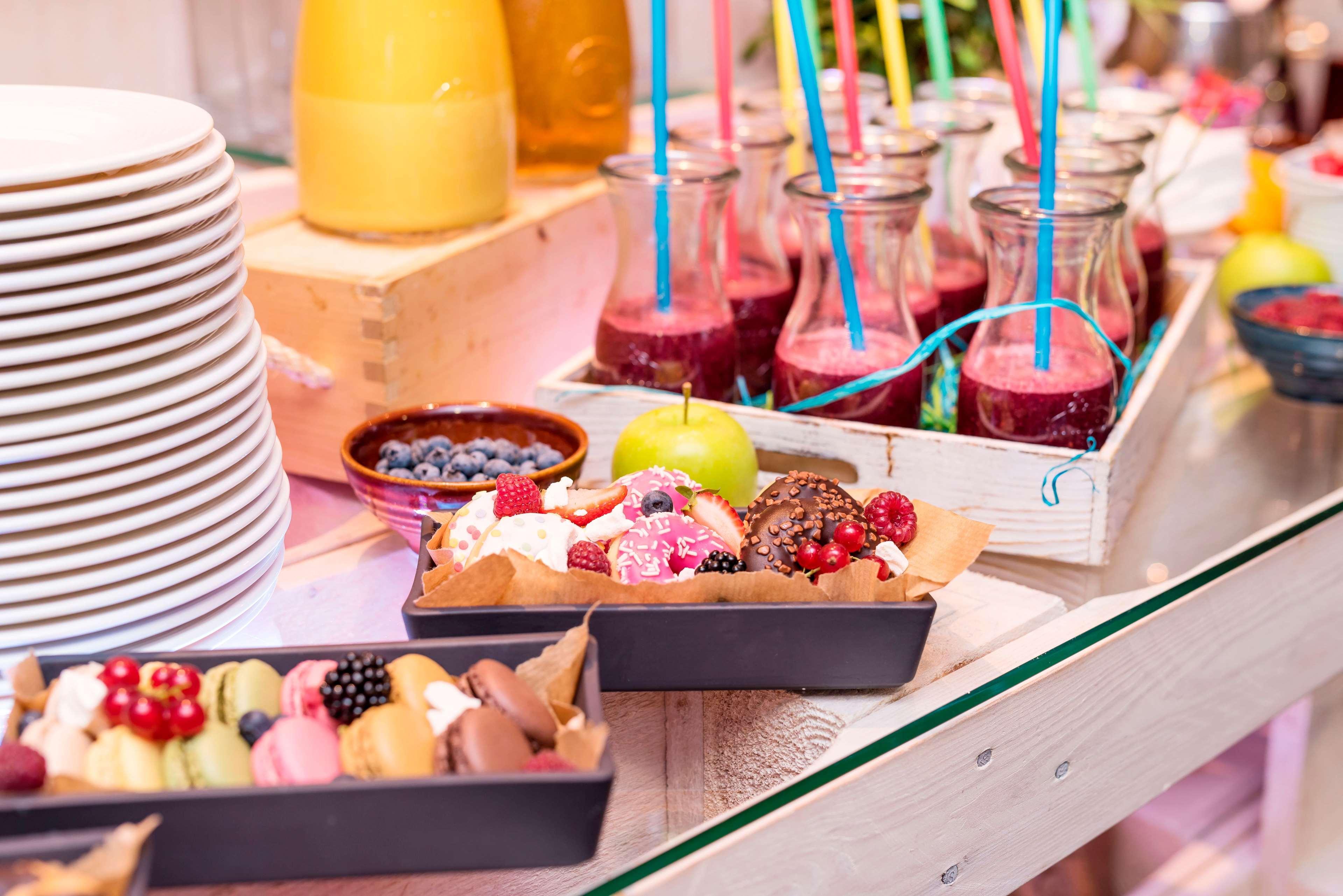 Radisson Blu Hotel Cracovia Esterno foto The photo features a vibrant display of desserts and drinks on a table. There are several stacked white plates on the left side. In the center, there are small glass bottles filled with a colorful smoothie or juice, each featuring a straw. A white wo