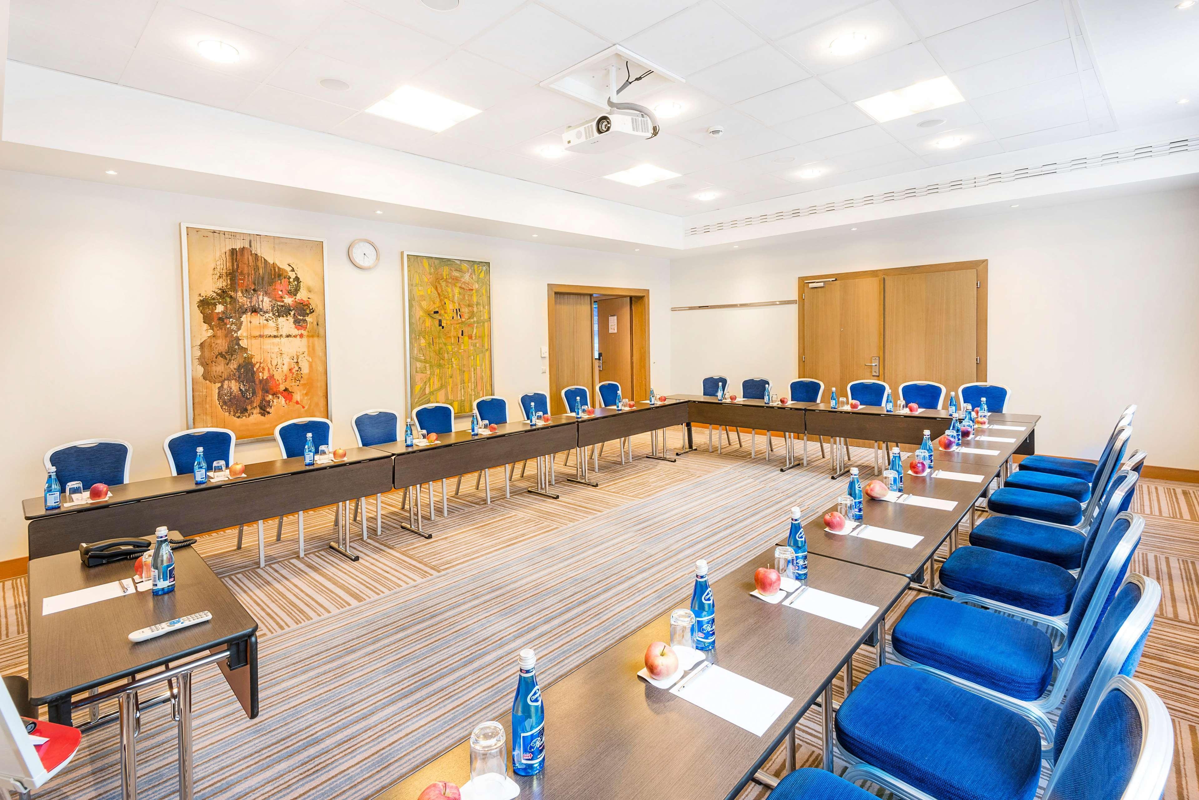 Radisson Blu Hotel Cracovia Esterno foto The photo shows a meeting room set up for a conference or workshop. There are two long tables arranged in a U-shape, with blue chairs positioned around them. Each table is equipped with bottles of water and apples, suggesting refreshments for attende