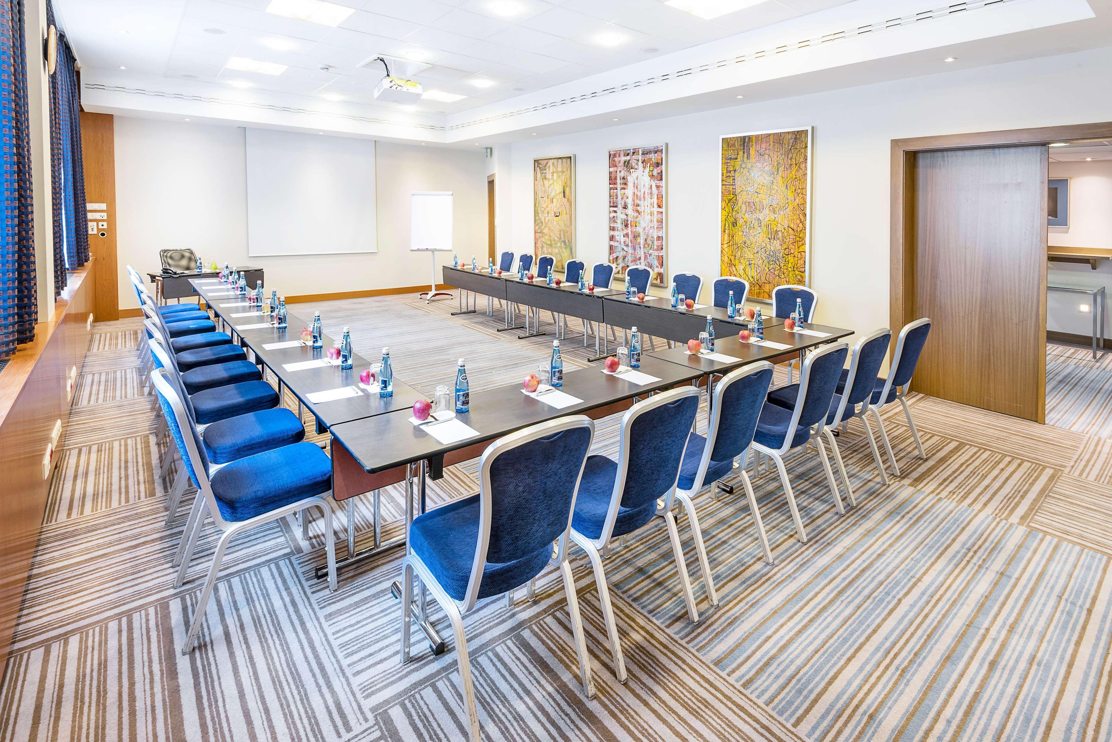 Radisson Blu Hotel Cracovia Esterno foto The image shows a conference room set up for a meeting. There are several long tables arranged in a U-shape, with blue chairs placed around them. Each chair has a small water bottle on the table, along with some stationery items, likely for the atten
