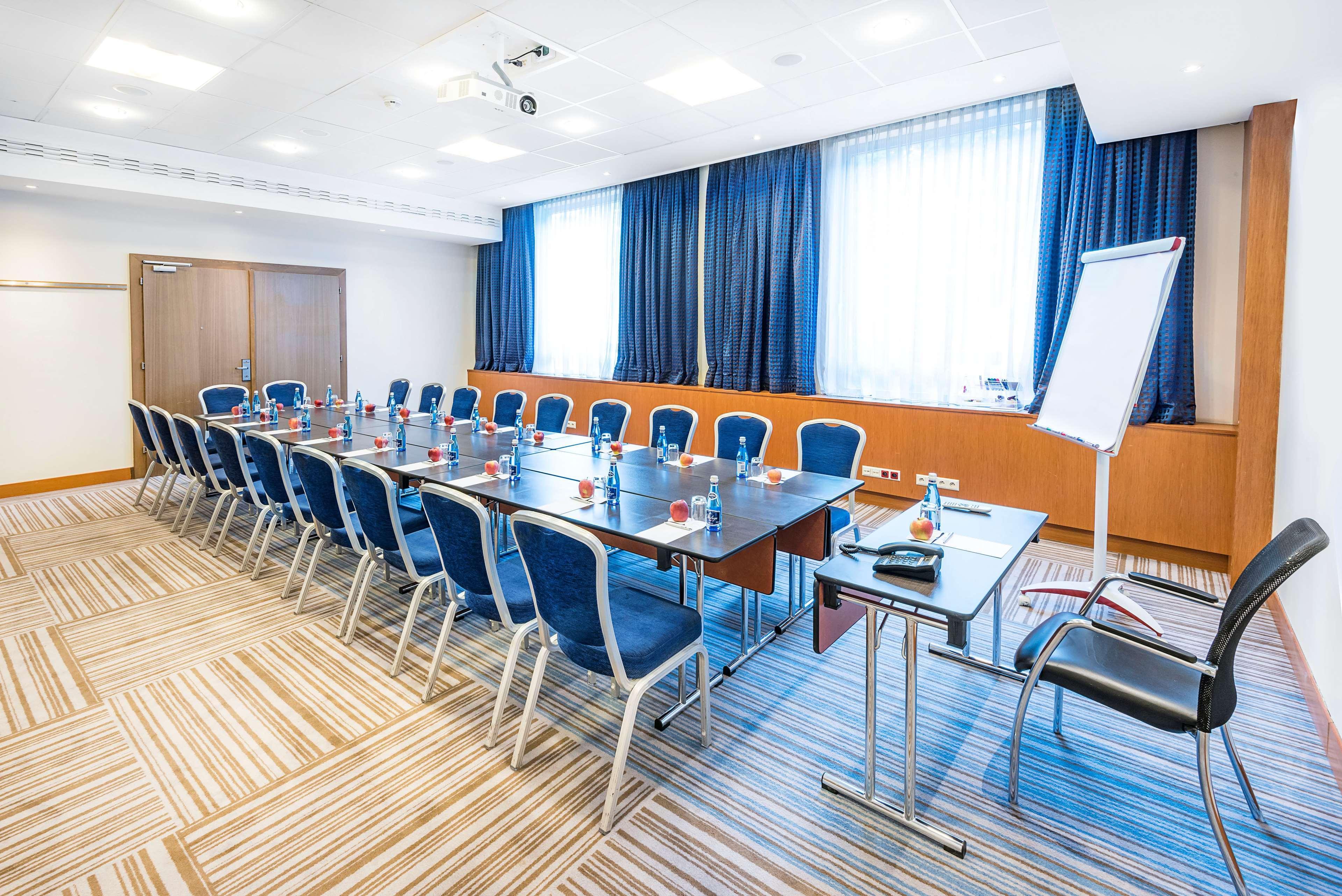 Radisson Blu Hotel Cracovia Esterno foto The photo shows a conference room set up for a meeting. There are several rows of blue chairs arranged around a long rectangular table. The table is equipped with small bottles of water, and there appears to be a flip chart or whiteboard on one side 