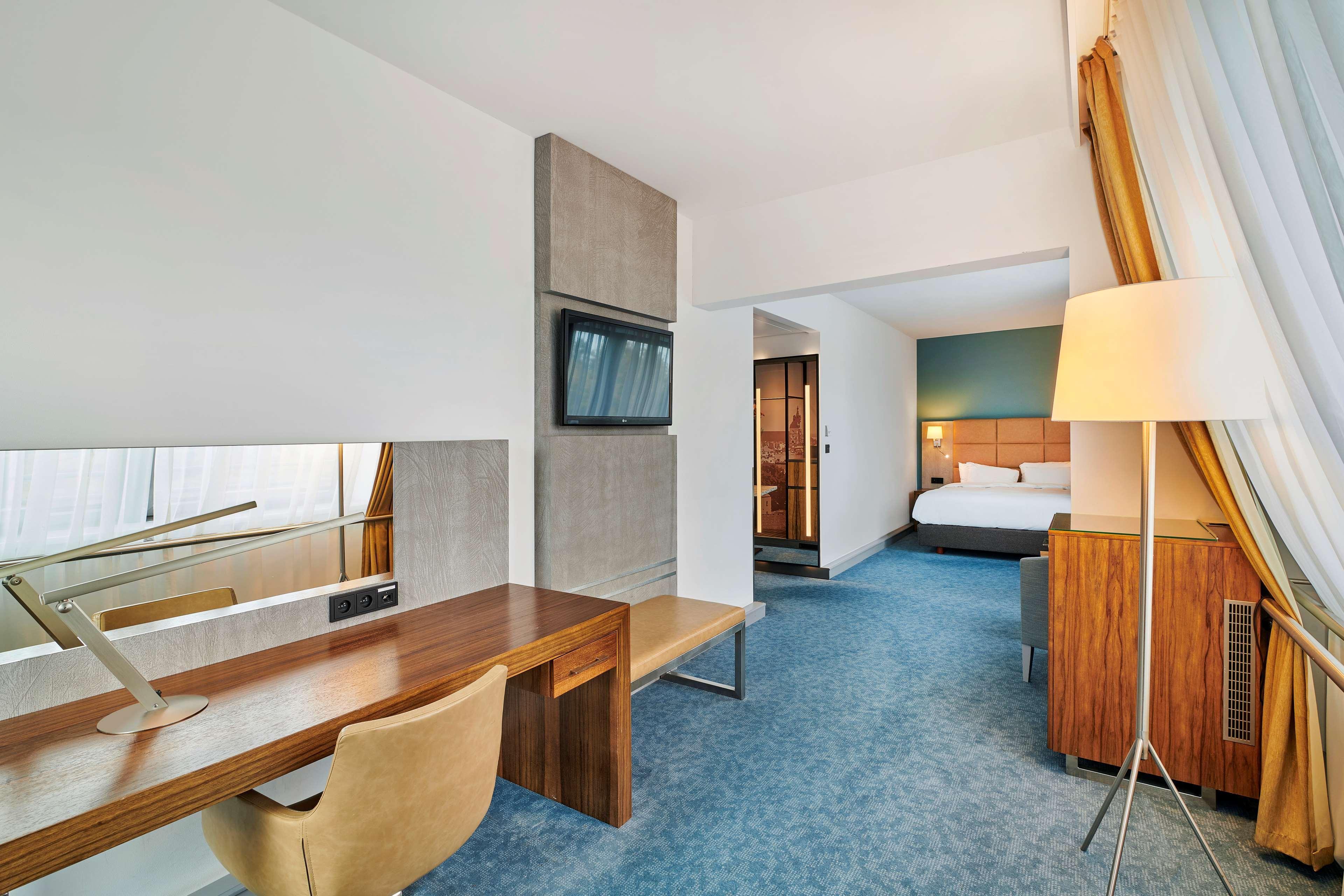 Radisson Blu Hotel Cracovia Esterno foto The photo showcases a modern hotel room. On one side, there is a work desk made of wood with a stylish chair. The desk is accompanied by a large mirror and a wall-mounted TV above it. The flooring is carpeted in a soft blue color. In the background, 