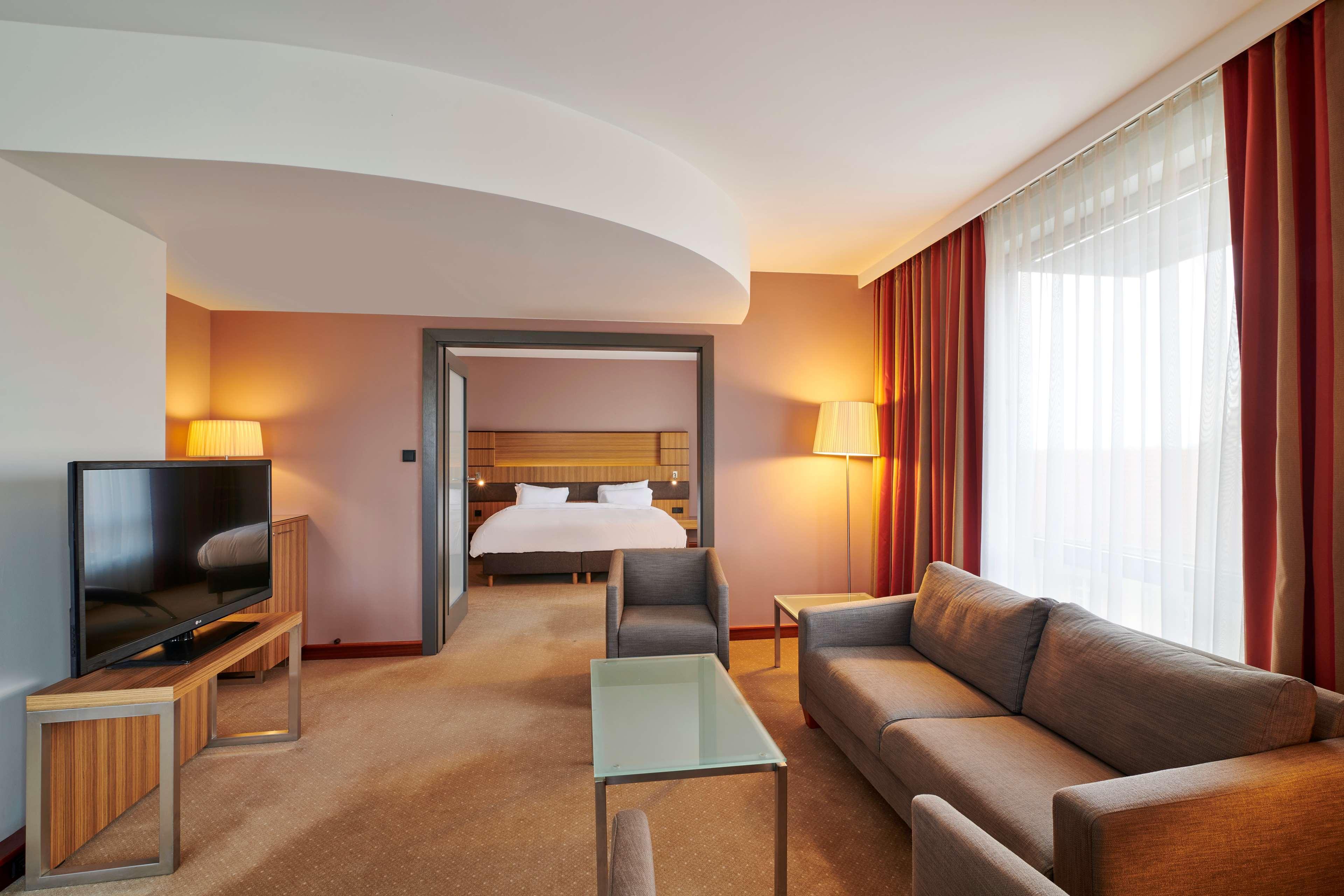Radisson Blu Hotel Cracovia Esterno foto The photo shows a modern hotel room suite. It features a cozy living area with a gray sofa, a glass coffee table, and a wooden TV stand with a flat-screen television. There are two lamps providing soft lighting. In the background, a door leads to the