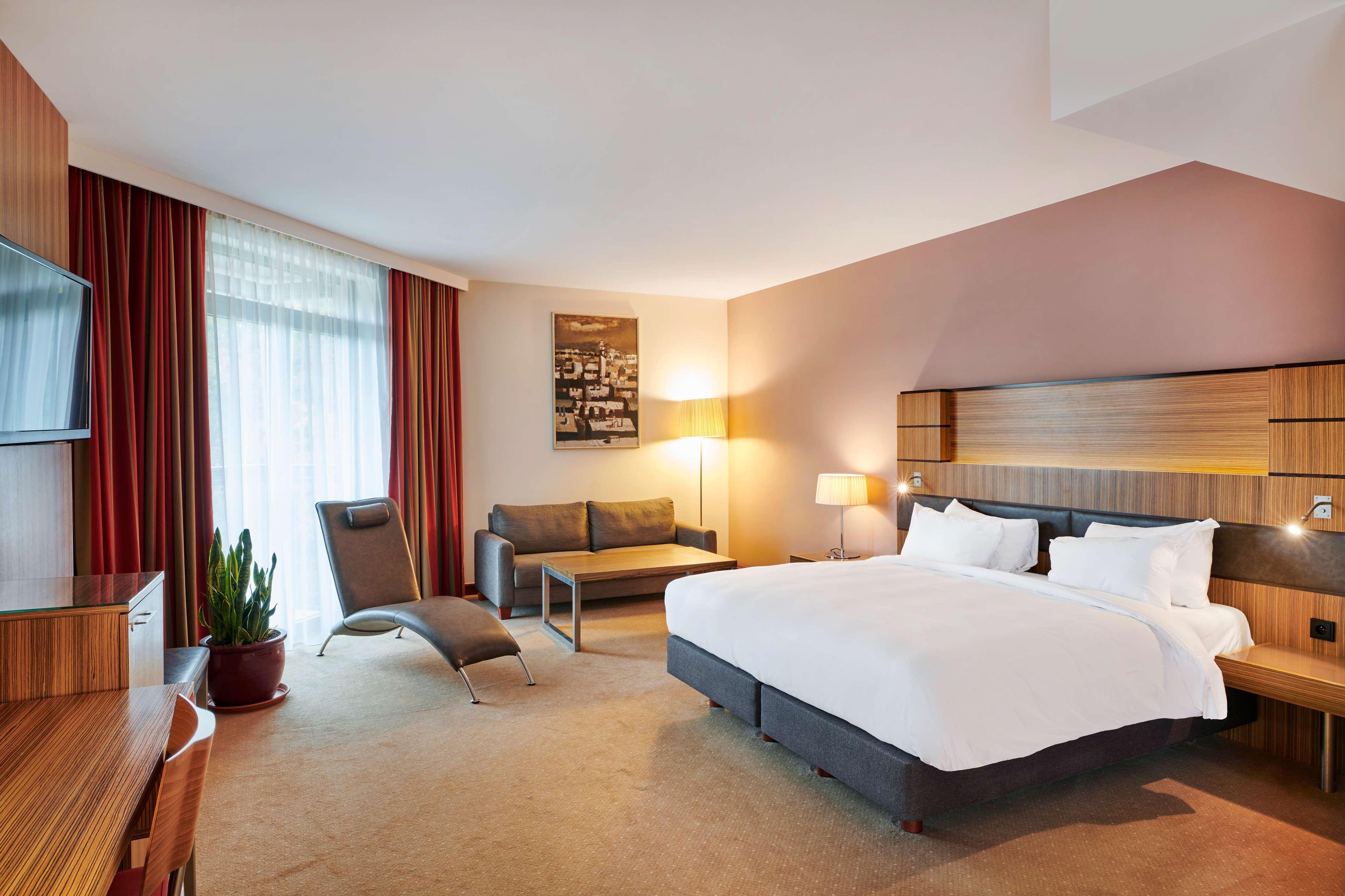 Radisson Blu Hotel Cracovia Esterno foto The photo shows a modern hotel room featuring a spacious layout. There is a large bed with white bedding positioned against a wooden headboard. Next to the bed, there is a small bedside table with a lamp. A couch is situated near a window dressed wit