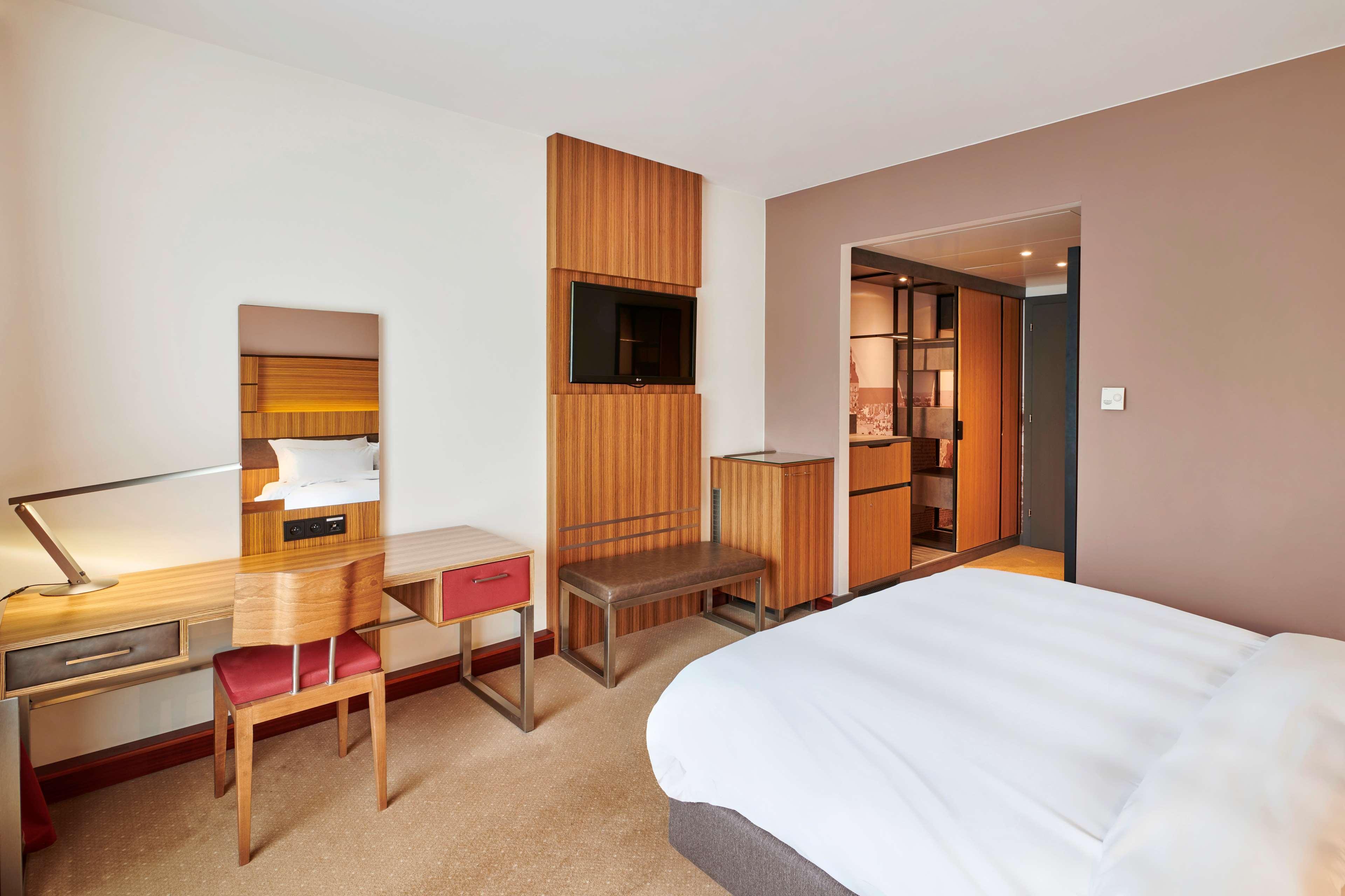 Radisson Blu Hotel Cracovia Esterno foto The photo shows a modern hotel room with a minimalist design. It features a large bed with white bedding, positioned against a wall. To one side, there's a small seating area with a simple bench. Opposite the bed is a desk with a mirror and a chair, 