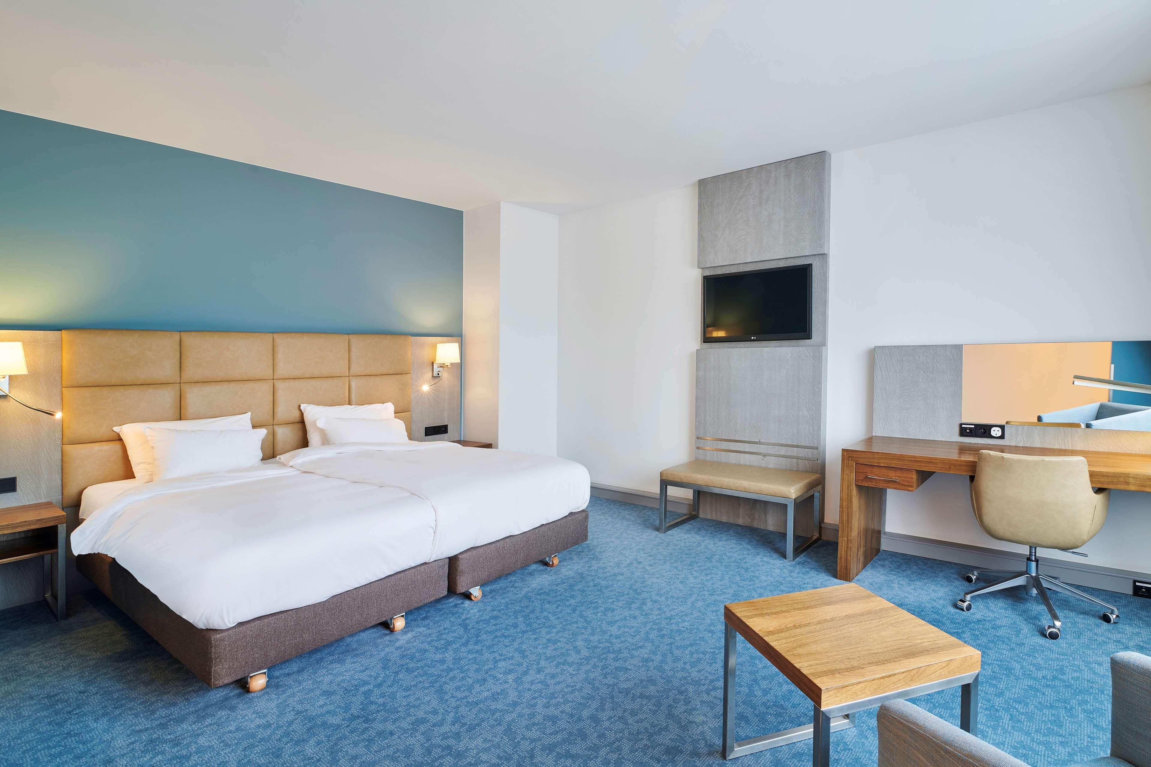 Radisson Blu Hotel Cracovia Esterno foto The photo shows a modern hotel room. It features two twin beds with white bedding and a light wooden headboard. The room has a light blue accent wall and a large window providing natural light. There is a flat-screen TV mounted on the wall opposite t