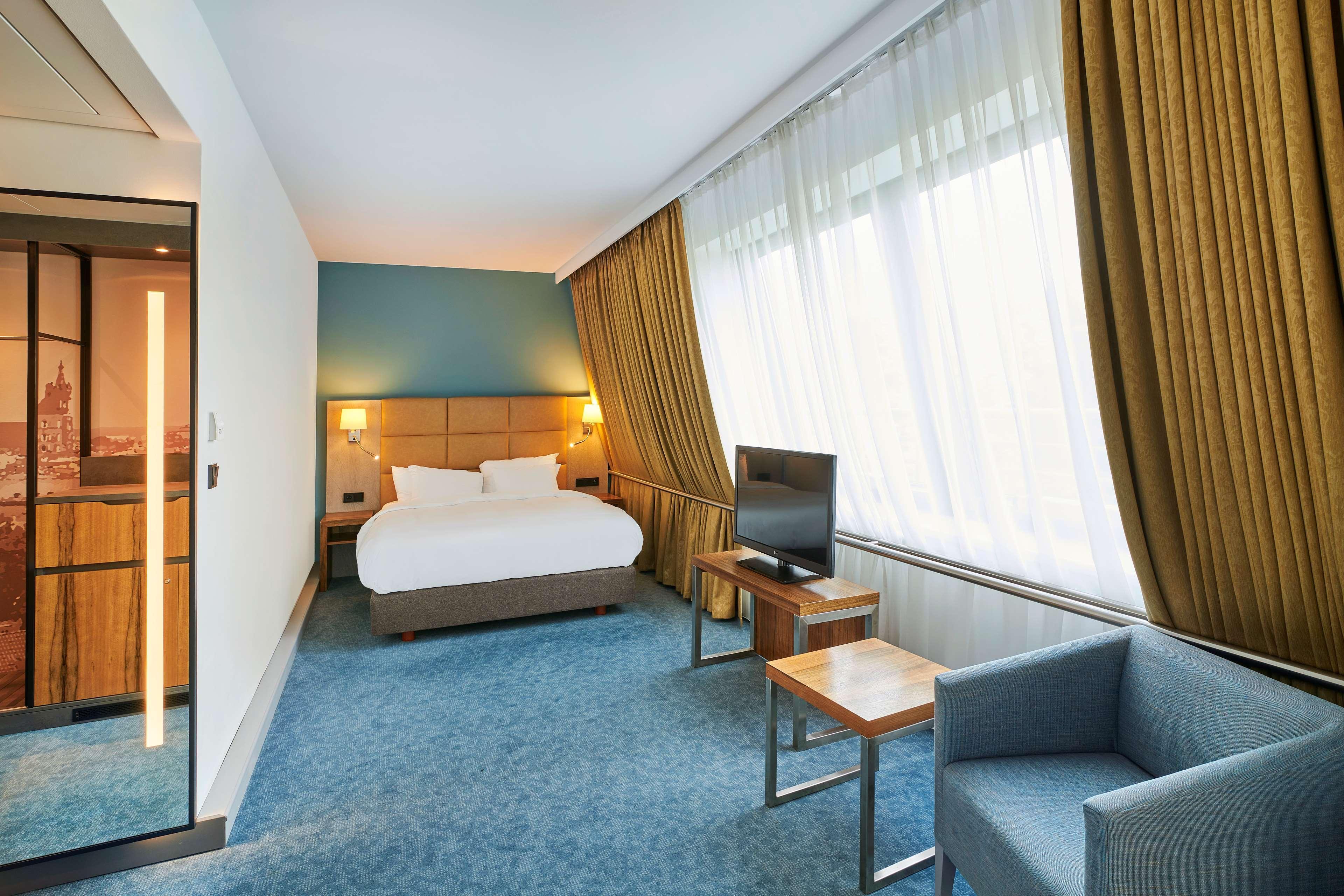 Radisson Blu Hotel Cracovia Esterno foto The photo shows a modern hotel room. It features a queen-sized bed with white bedding, positioned against a wall with warm lighting. The room has a large window with sheer curtains that allow natural light to enter. There's a small wooden table and a