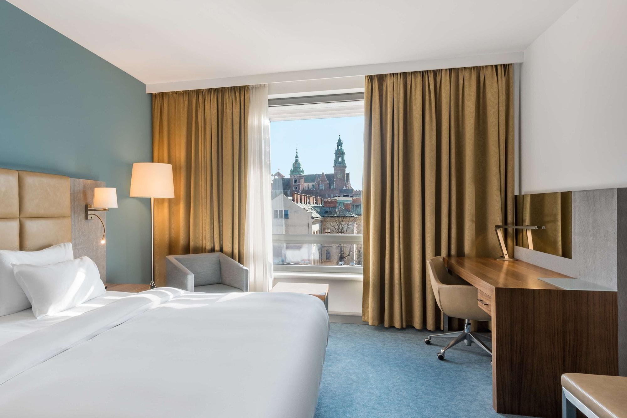 Radisson Blu Hotel Cracovia Esterno foto The photo depicts a hotel room featuring a large bed with white linens, a light-colored headboard, and a side lamp. There is a comfortable armchair in the corner, and a wooden desk with a chair positioned near the window. The window reveals a view of