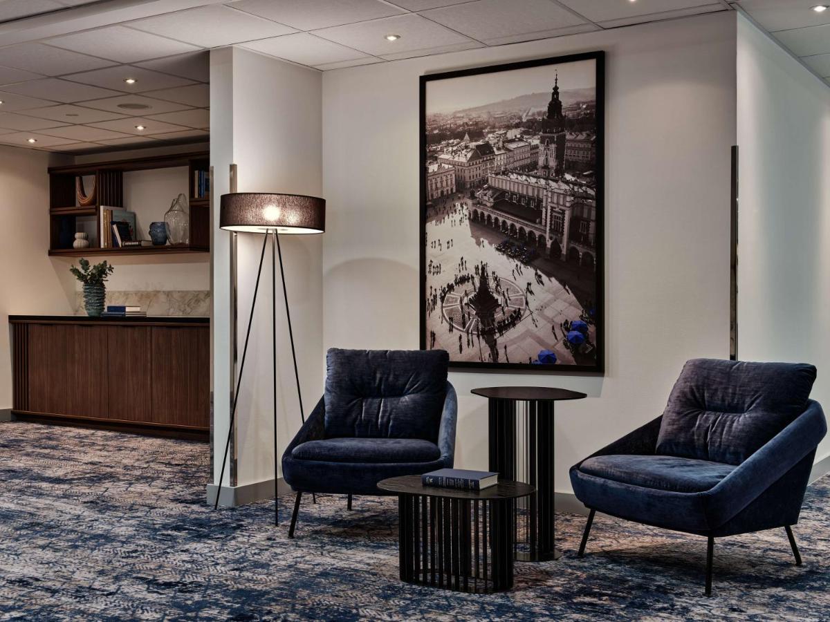 Radisson Blu Hotel Cracovia Esterno foto The photo depicts a modern interior space that appears to be part of a hotel or a lounge area. In the foreground, there are two stylish blue armchairs accompanied by a small round black table. To the left, there is a reception desk with a decorative 