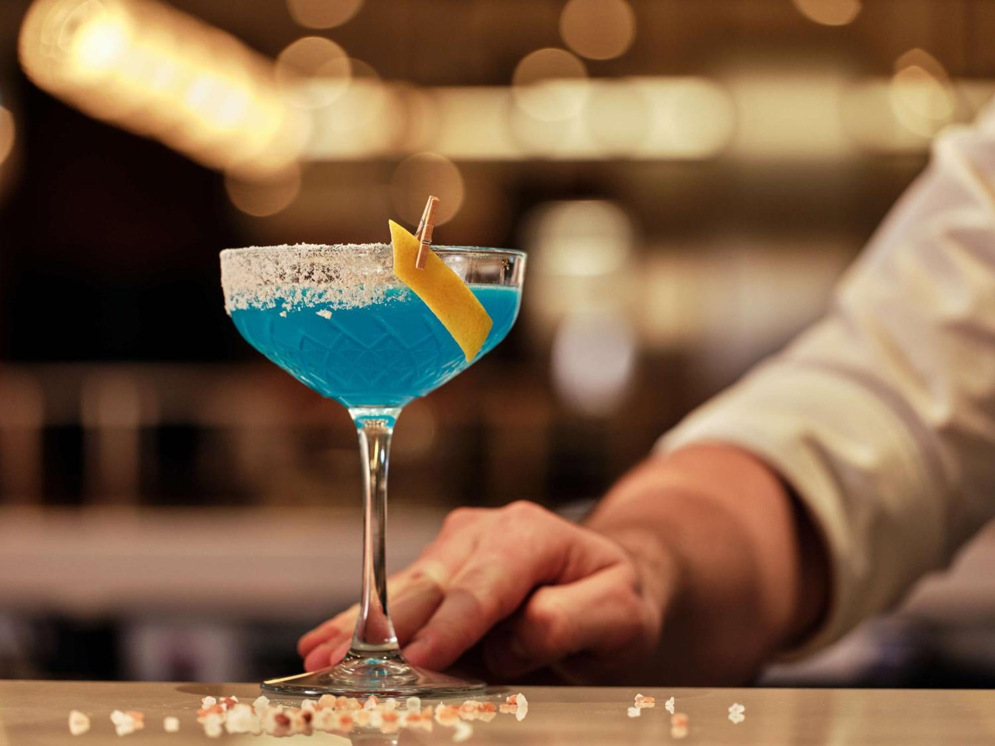 Radisson Blu Hotel Cracovia Esterno foto The photo shows a vibrant blue cocktail served in a classic cocktail glass. The drink is garnished with a slice of lemon and has a rim coated with salt. In the background, there are blurred lights, creating a lively atmosphere, and a hand is gently p