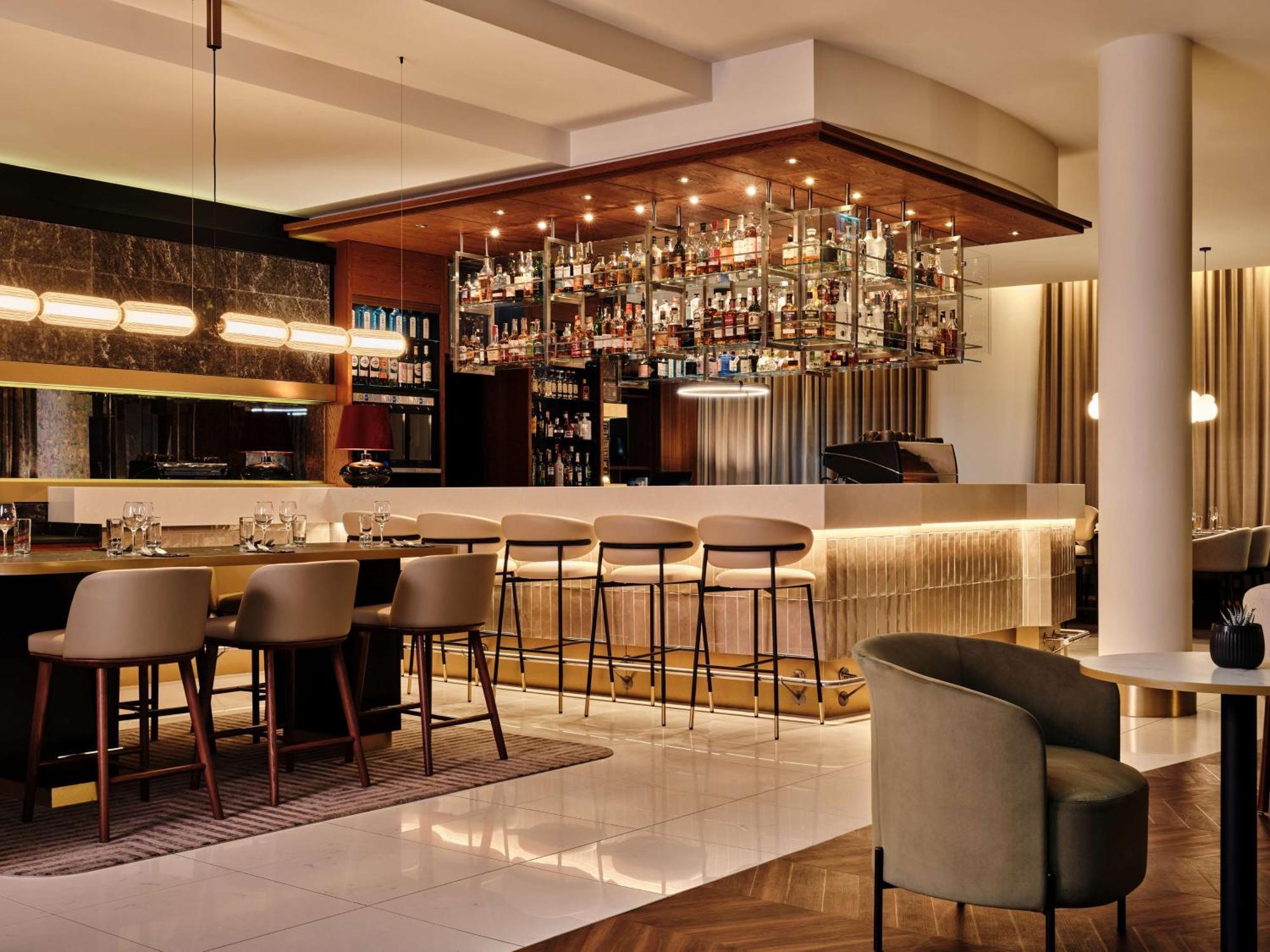 Radisson Blu Hotel Cracovia Esterno foto The image shows a stylish bar area. There is a modern bar with a sleek design featuring a long counter and high stools. Above the bar, there is a display of various bottles, arranged neatly on shelves, illuminated by soft lighting. The overall ambian