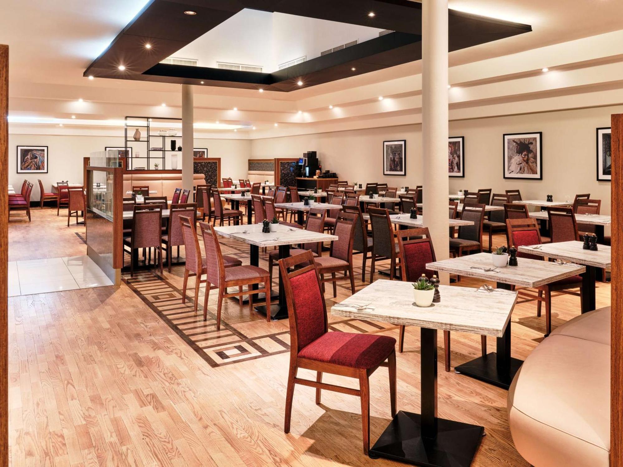 Radisson Blu Hotel Cracovia Esterno foto The photo shows a spacious and modern restaurant interior. It features multiple tables surrounded by wooden chairs, some of which have red upholstery. The floor is made of light-colored wood, and there's a geometric pattern visible in the design. The