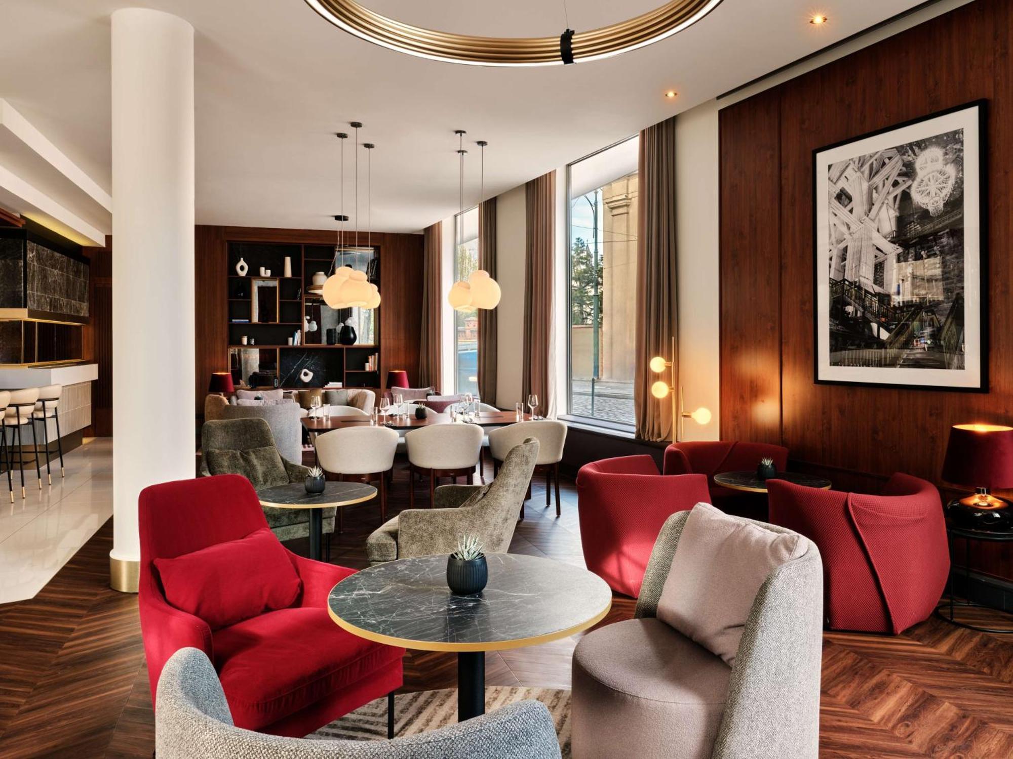 Radisson Blu Hotel Cracovia Esterno foto The image depicts a stylish, modern interior of a lounge or café. The space features a mix of seating options, including red and gray armchairs arranged around small circular tables. The décor is warm and inviting, with wooden paneling on the walls a