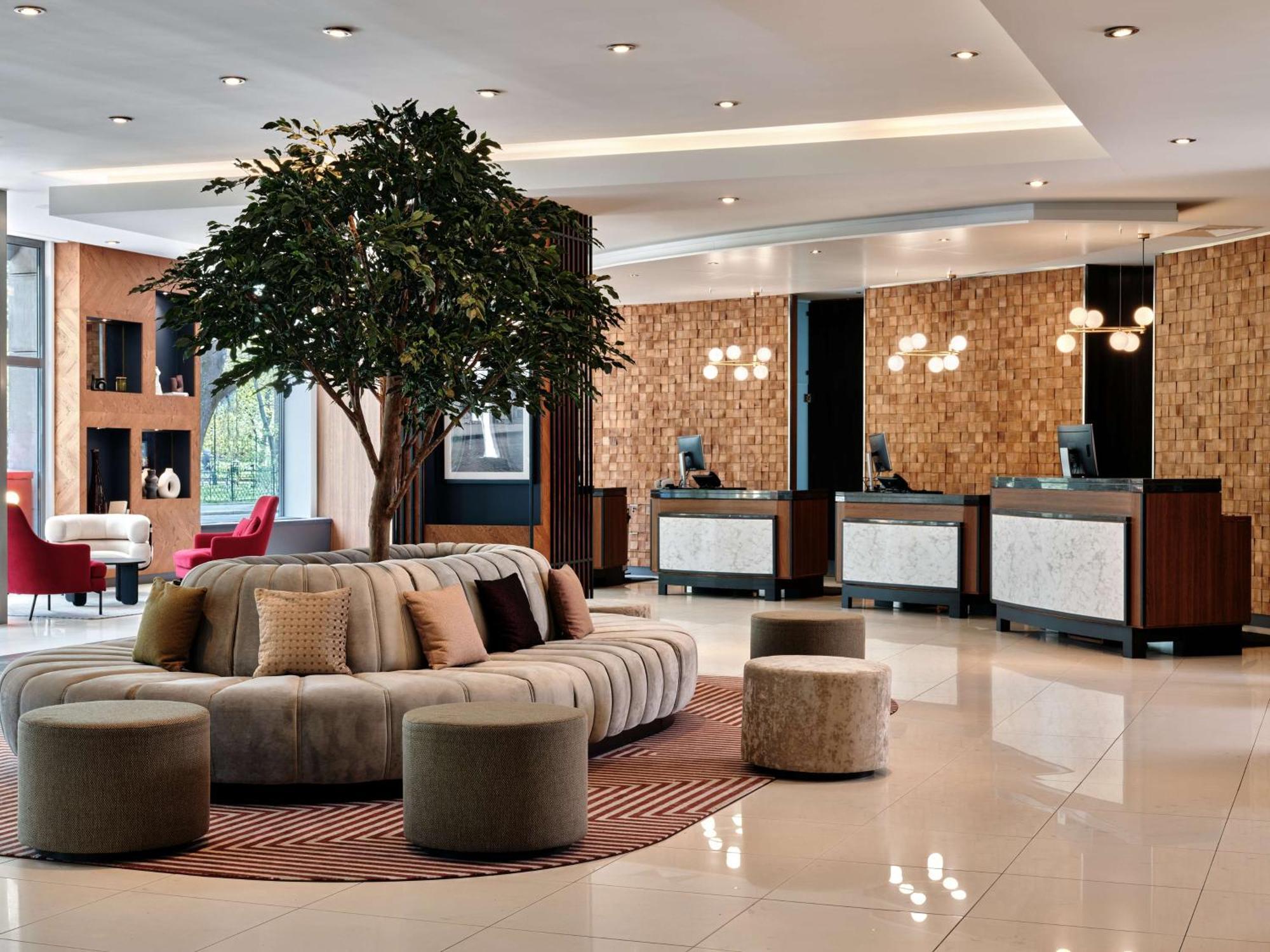 Radisson Blu Hotel Cracovia Esterno foto The photo shows a modern hotel lobby. The space features a circular, plush seating area in neutral tones, surrounded by small, round stools. A large decorative tree adds a touch of greenery to the room. In the background, there are multiple reception