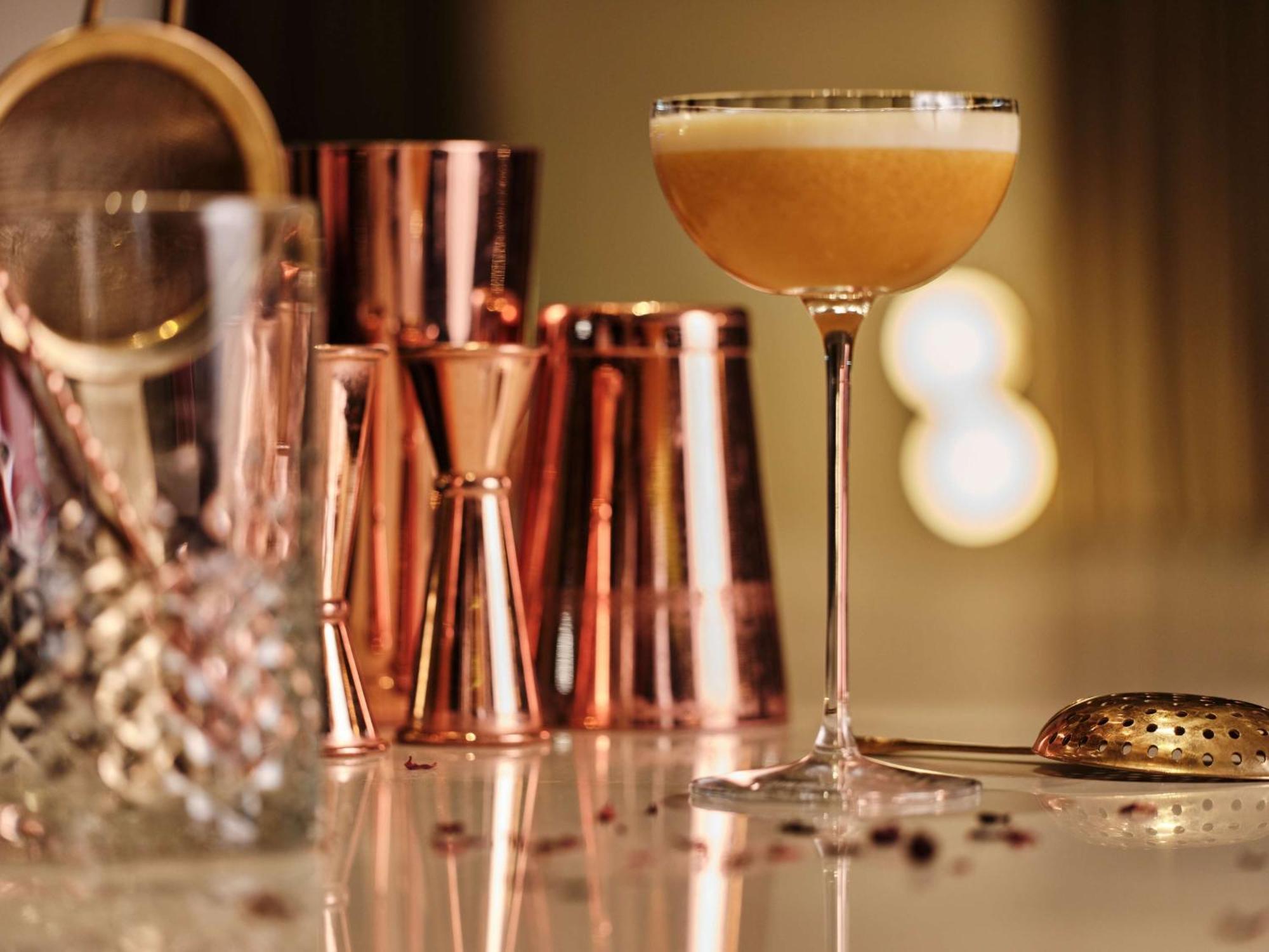Radisson Blu Hotel Cracovia Esterno foto The image shows a stylish cocktail setup. In the foreground, there is a beautifully crafted cocktail glass filled with a light-colored drink topped with a frothy layer. Next to it, you can see several copper-colored bar tools, including jiggers (meas