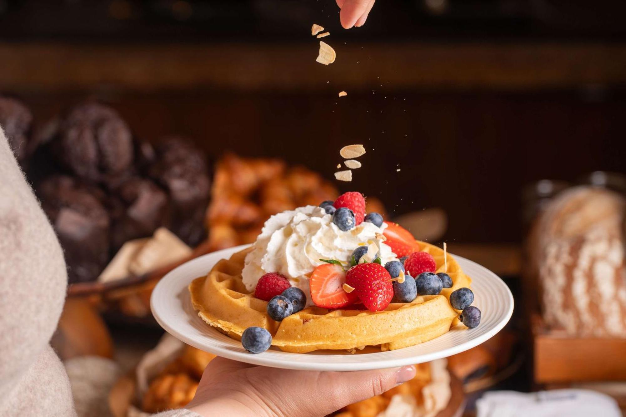 Radisson Blu Hotel Cracovia Esterno foto The photo features a hand sprinkling nuts or some form of topping over a plate of waffles that is beautifully garnished with whipped cream, strawberries, raspberries, and blueberries. In the background, there are various baked goods, likely indicatin