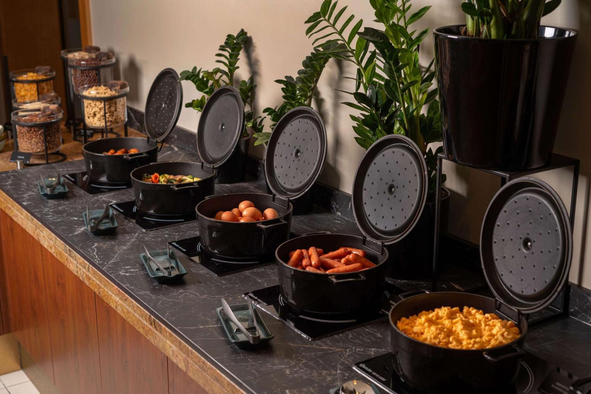 Radisson Blu Hotel Cracovia Esterno foto The photo shows a buffet setup with several black cooking pots arranged in a row on a dark countertop. Each pot is covered with a lid, and some pots are filled with various prepared foods. The dishes visible include eggs, hot dogs, macaroni and chees