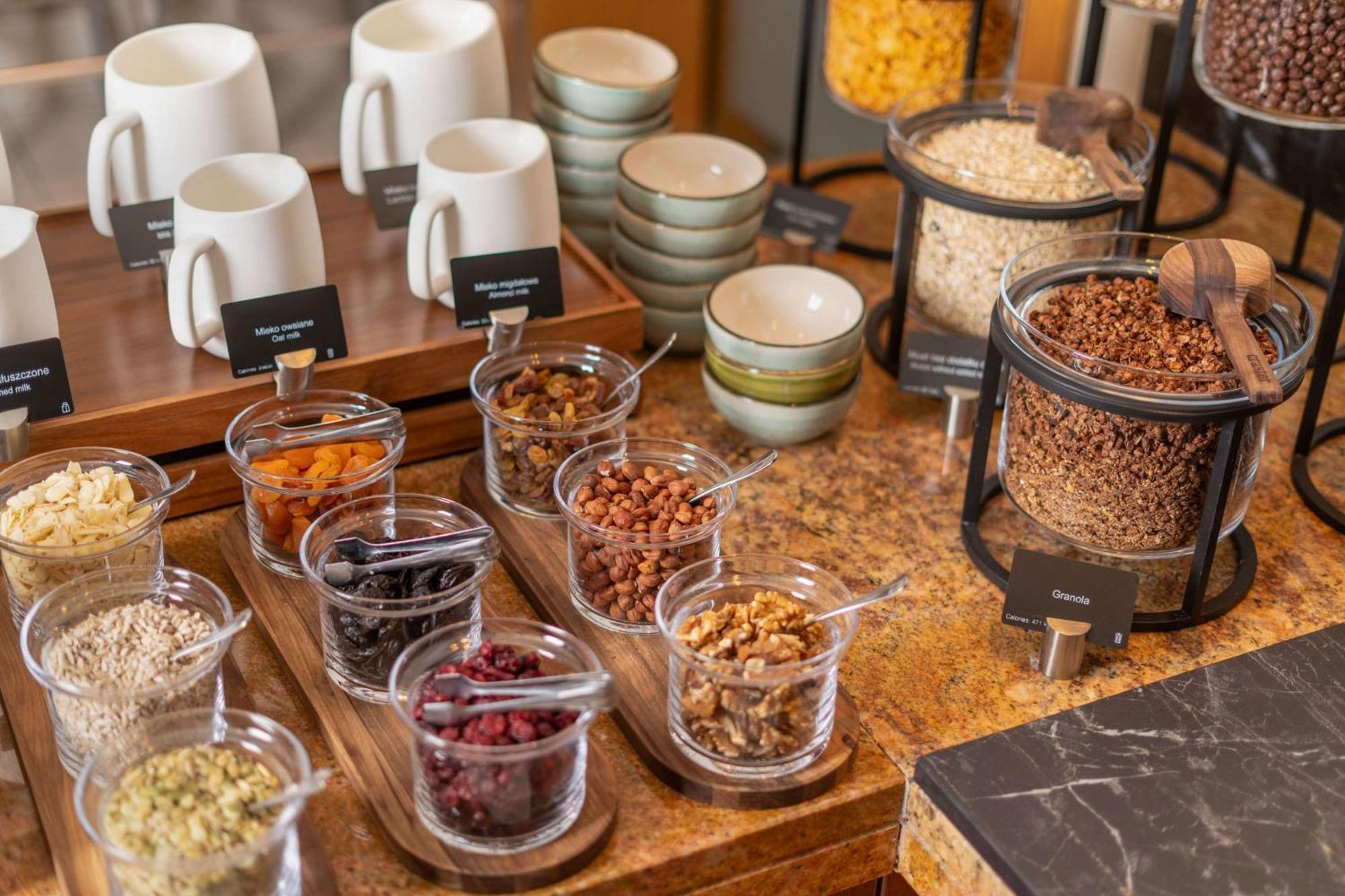 Radisson Blu Hotel Cracovia Esterno foto The photo displays a breakfast or snack buffet setup with an assortment of items. There are several white mugs and smaller bowls on a wooden tray, indicating that beverages may be served. In the foreground, there are various glass containers filled w